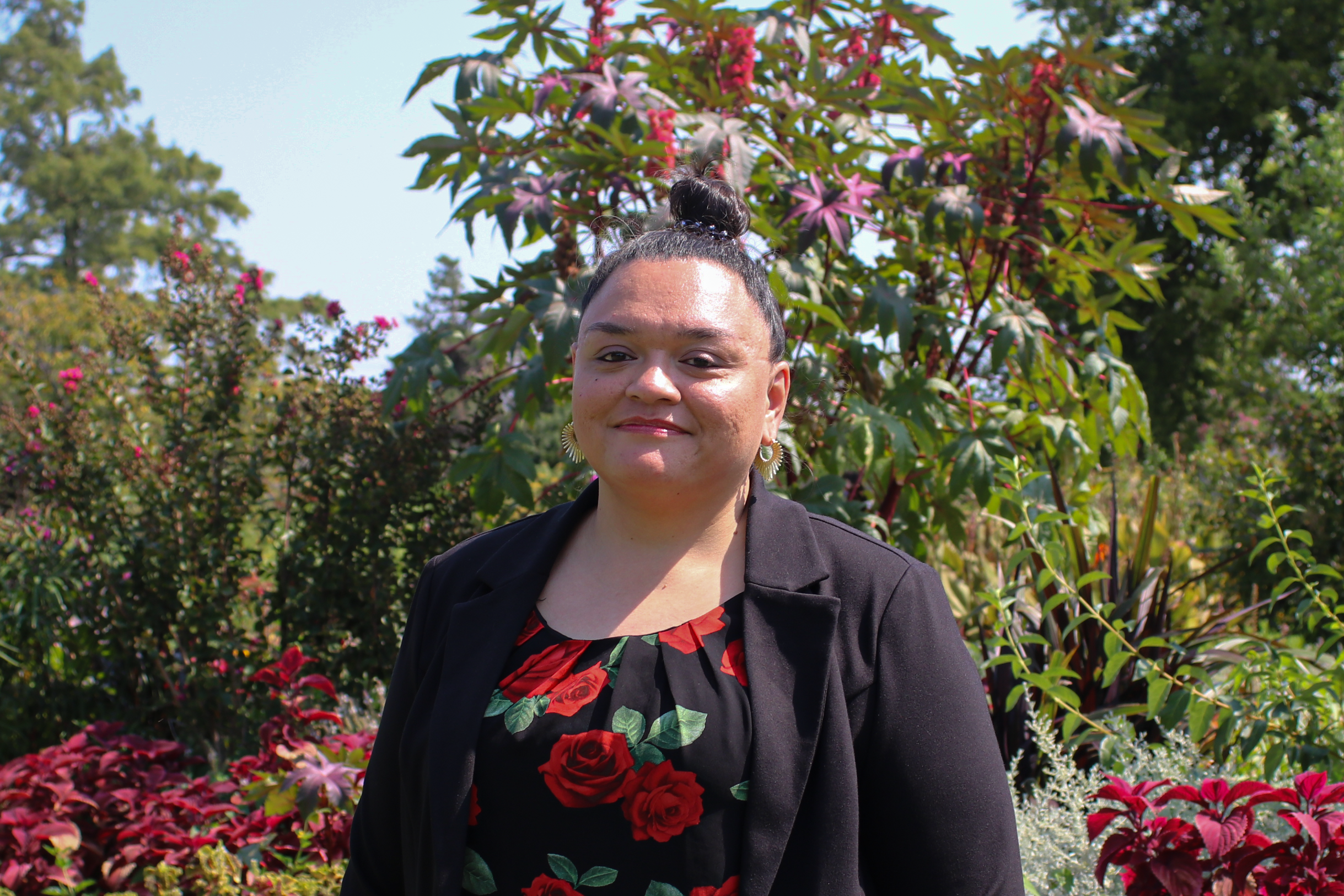 Smiling woman in black floral top w/ black blazer on a bright sunny day w/ a colorful garden behind her. Image © Kacie Puckett Photography