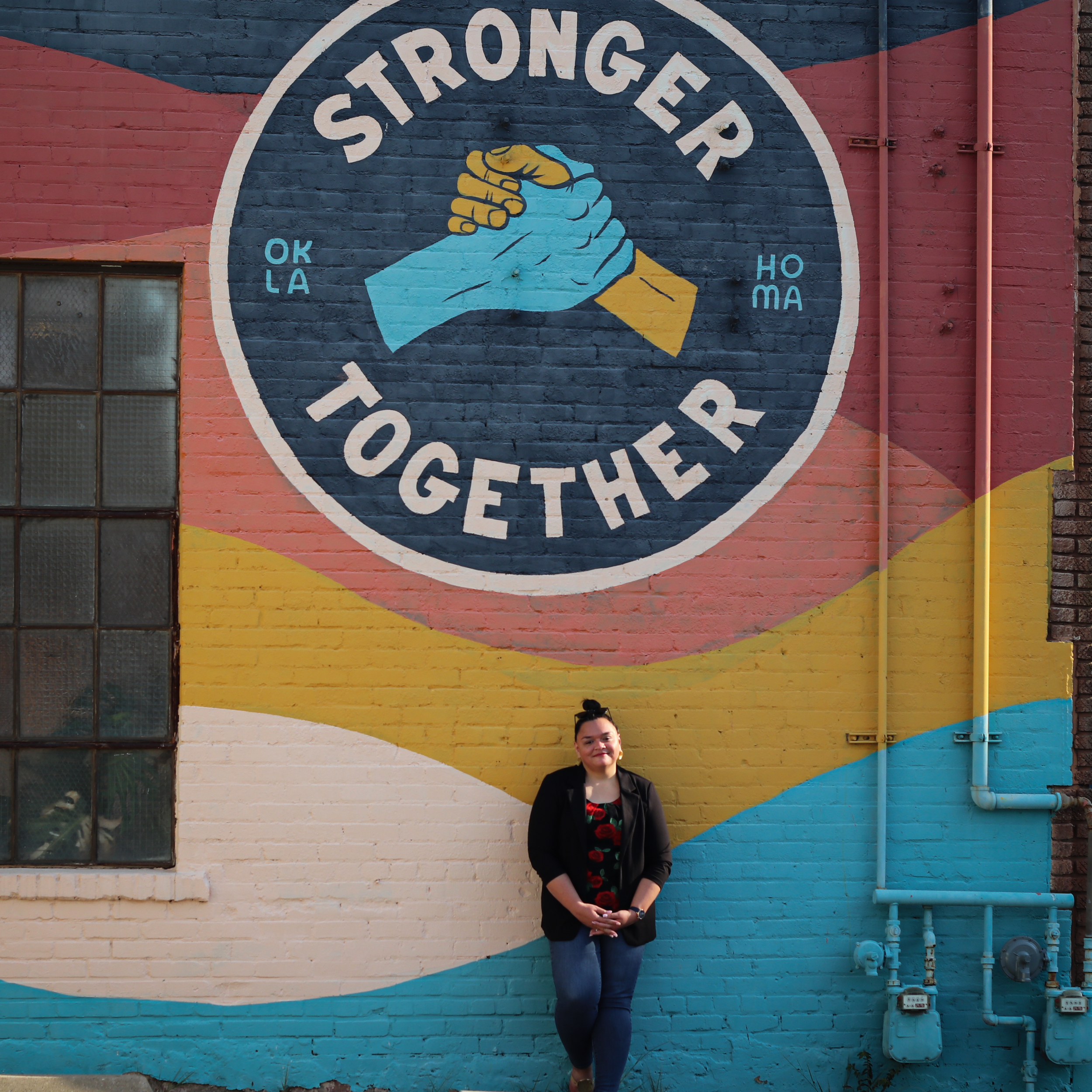 Smiling woman in business casual attire standing slightly off center under an Oklahoma-themed mural of two hands held in solidarity that reads " Stronger Together". Image © Kacie Puckett Photography