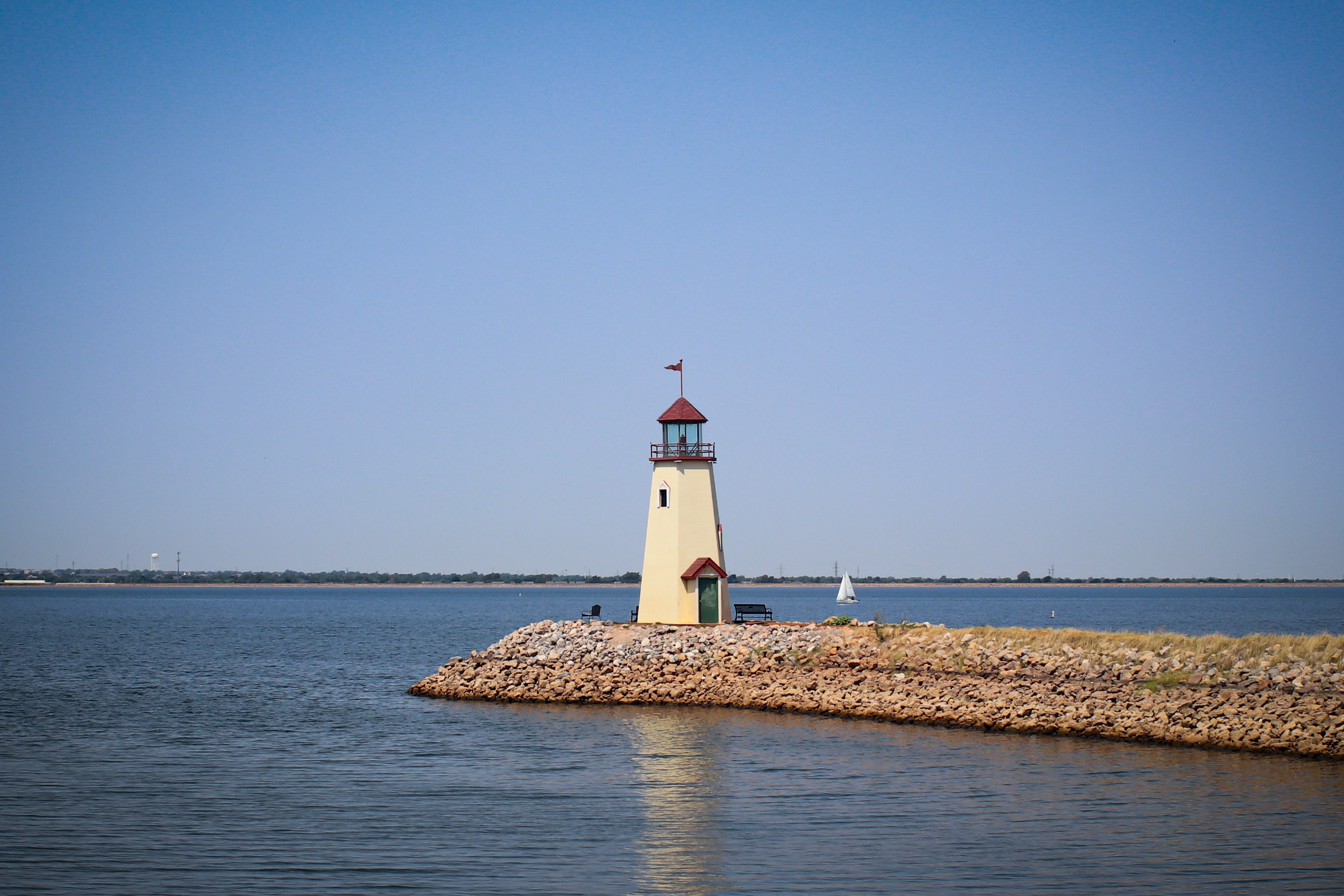 Lake Hefner Lighthouse in Oklahoma City on a bright, cloudless, sunny day with blue skies and a small white sailboat far in the background. Image © Kacie Puckett Photography