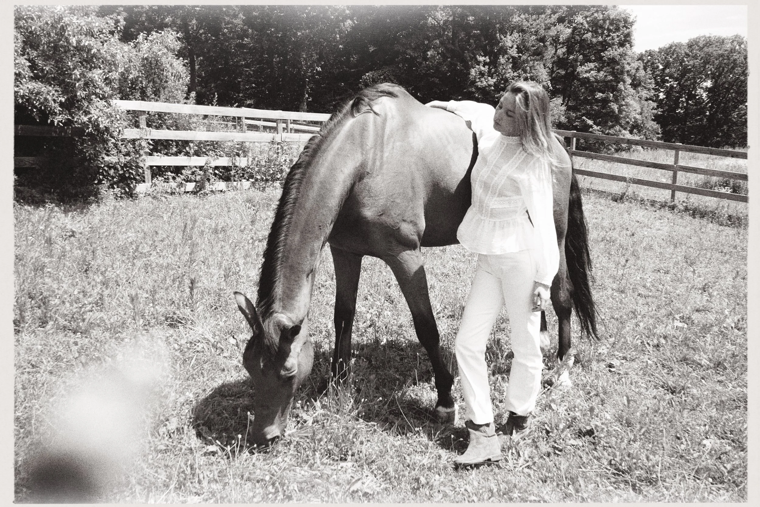 A woman with long hair and light-colored clothing standing outdoors, gently petting a horse near a wooden fence and trees.