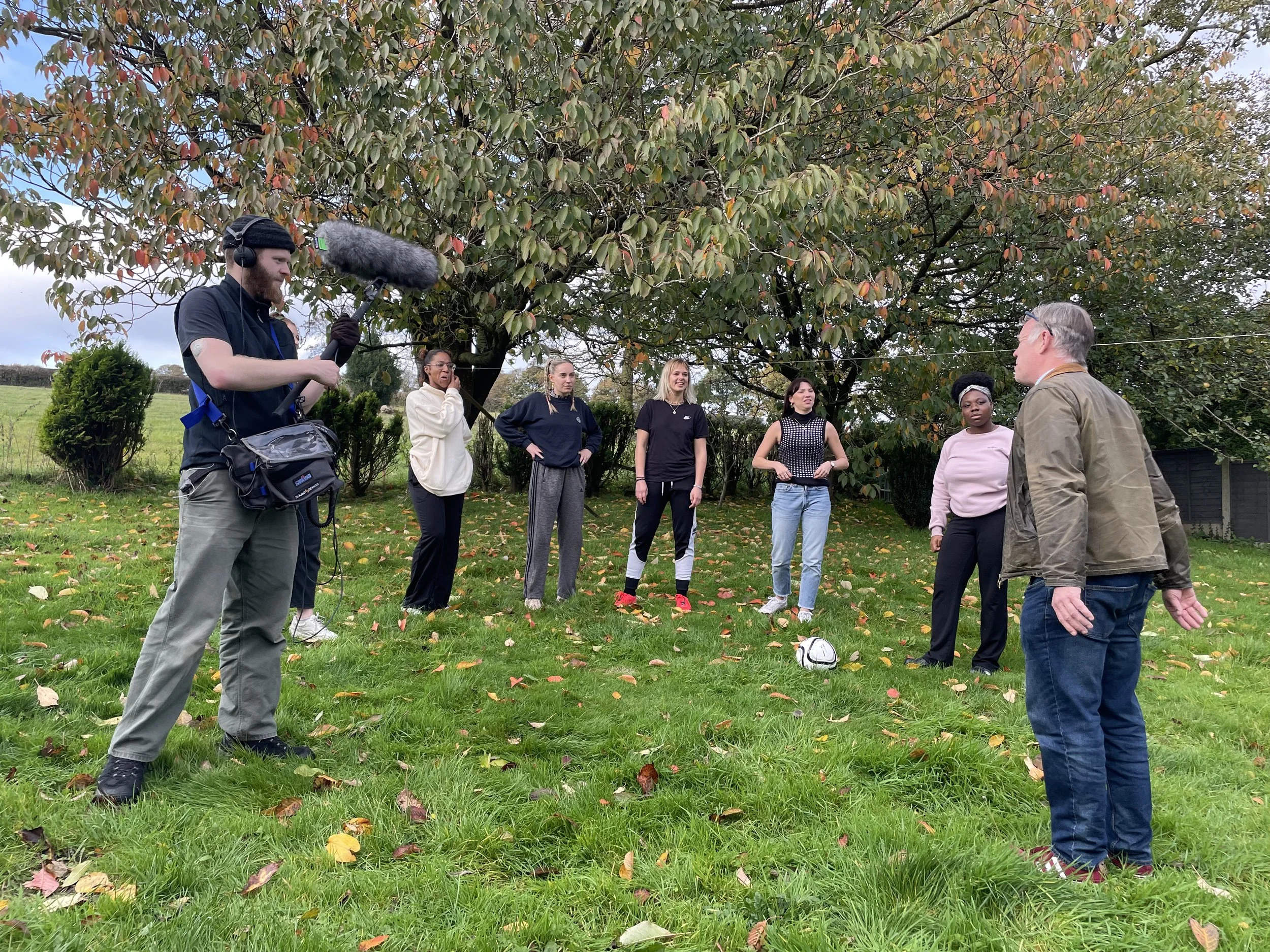 A group of six women and one man standing outdoors on grass with fallen leaves, under a tree with dense foliage. A man is talking to the group, and a man on the left is filming with a boom microphone. The scene appears to be a discussion or interview in a park or garden.
