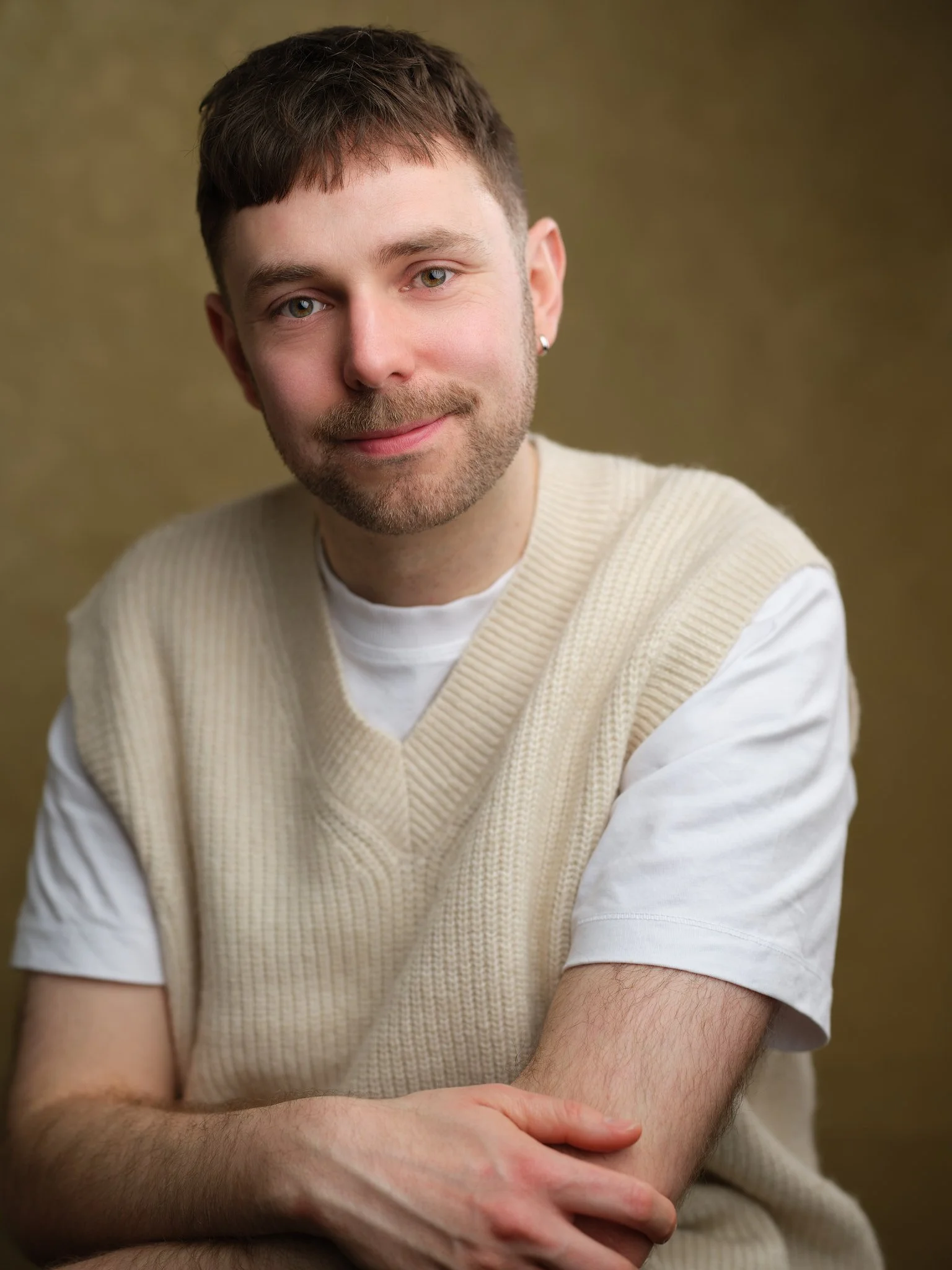 A young man with short brown hair, light facial hair, and green eyes, wearing a cream-colored sleeveless sweater over a white t-shirt, sitting with arms crossed and smiling softly against a brown background.