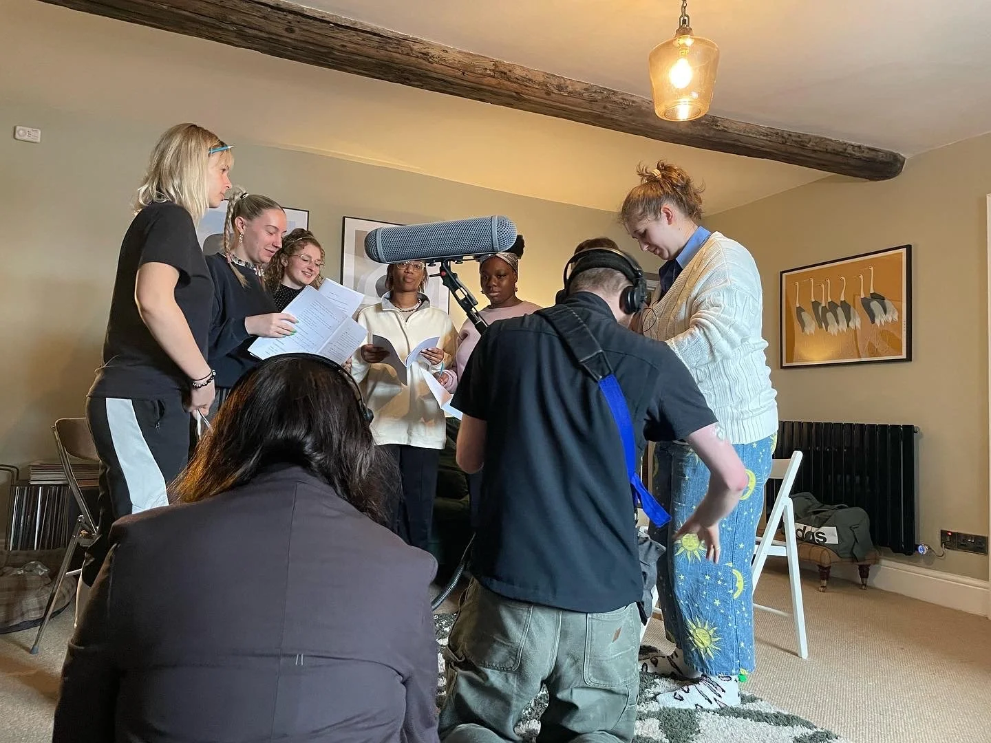 A group of people standing together in a room with one person recording with a camera and microphone, while others are reading from scripts or pages, possibly rehearsing or recording a scene.