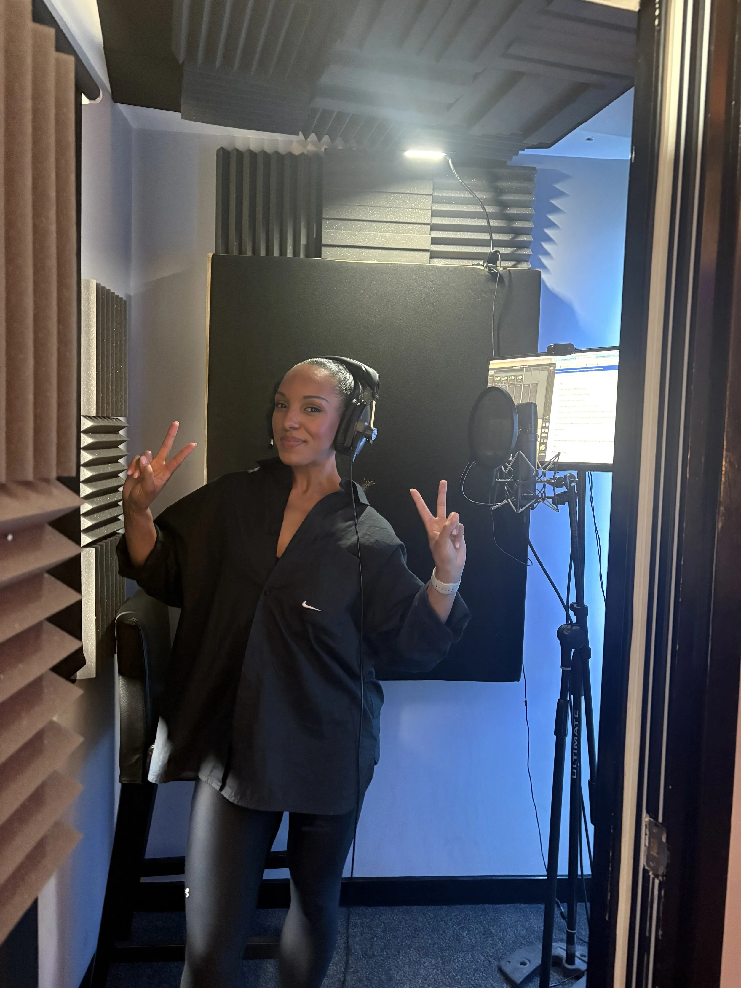 A woman inside a vocal booth, wearing headphones and making peace signs with both hands, surrounded by acoustic foam panels, with a computer and microphone in front of her.