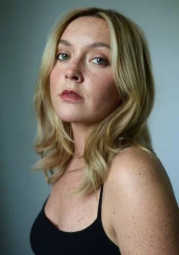 A young woman with blonde hair, light skin, and freckles, looking over her shoulder at the camera, wearing a black sleeveless top against a plain gray background.