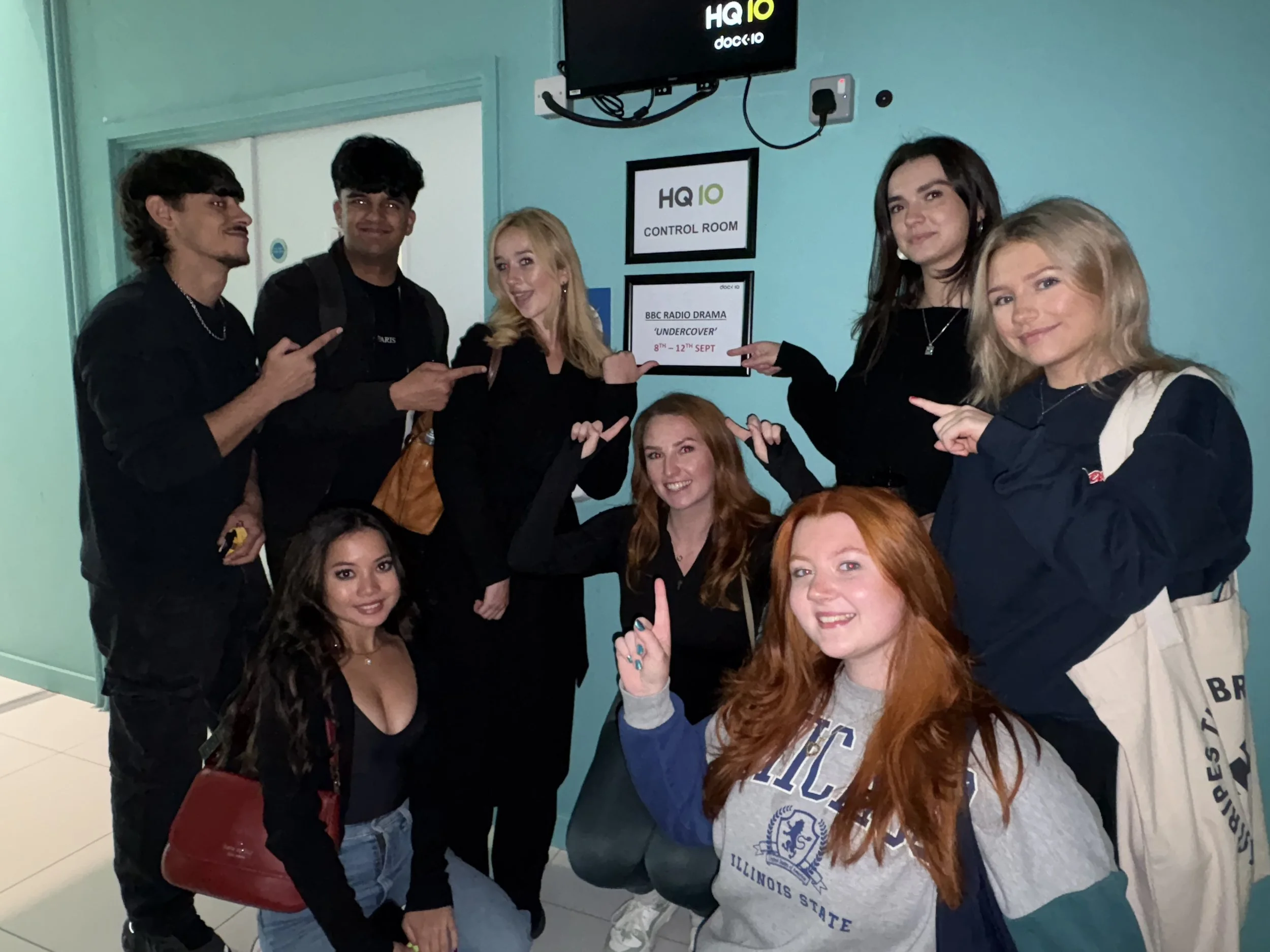 Group of six young adults, four women and two men, posing indoors in front of a wall with signs indicating HQ10 control room and BBC Radio Drama 'Undercover'. The women are smiling, one wearing a sweatshirt with Illinois State University logo, and the men are pointing at one woman.