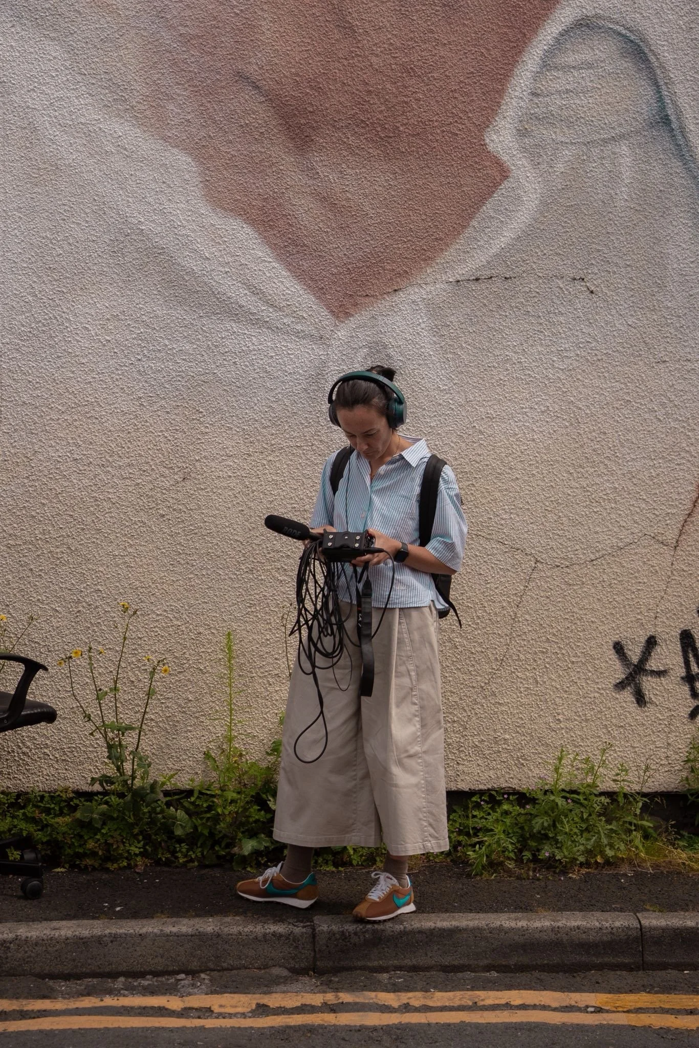 A woman in a light blue striped shirt, baggy beige pants, and sneakers with teal and brown accents is standing on a sidewalk, looking at a device in her hands. She is wearing headphones and has a black bag on her shoulder. Behind her is a beige textured wall with a faded mural of a heart shape, some small plants, and graffiti on the lower right corner of the wall.