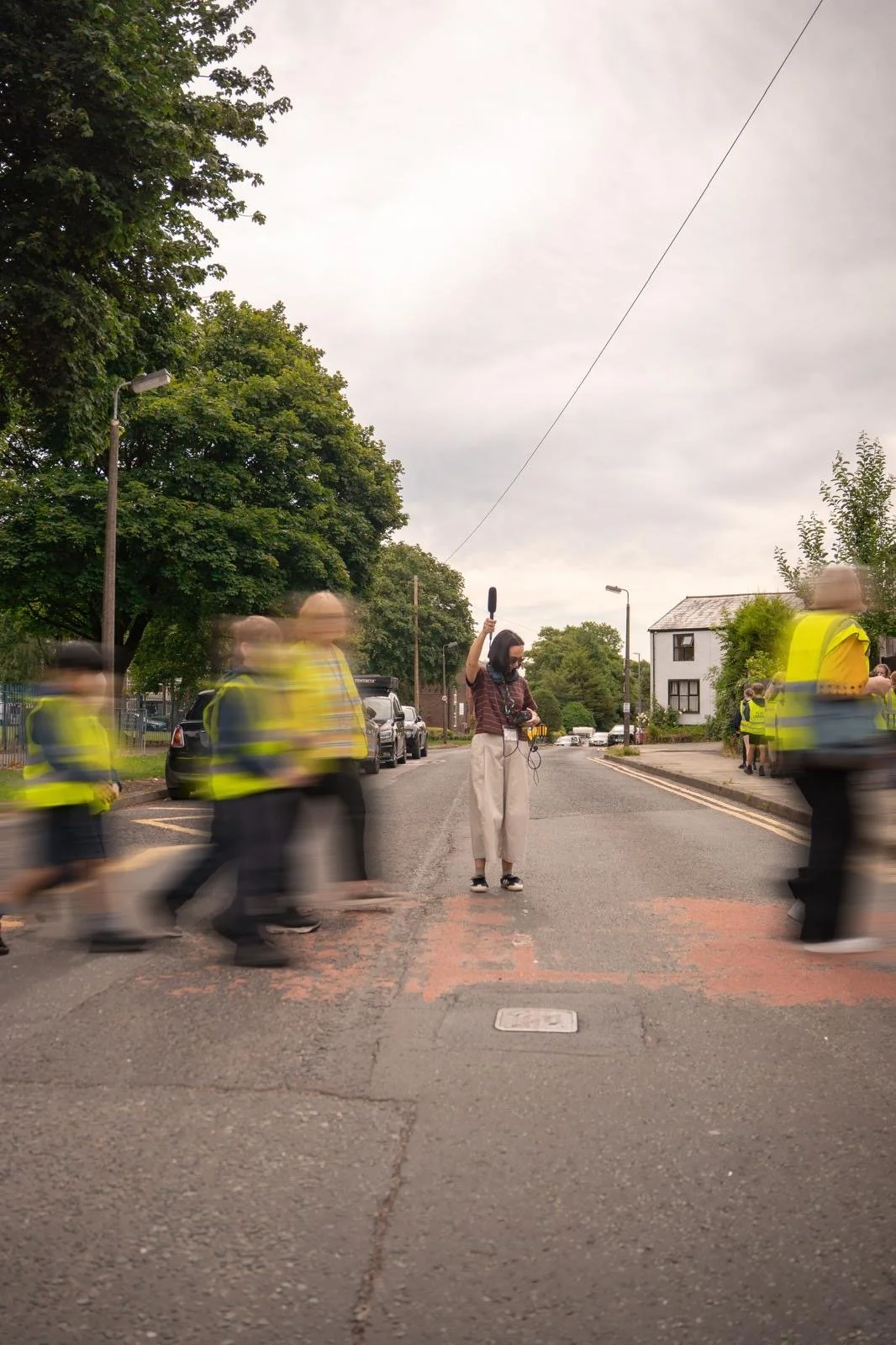 A woman with a microphone and headphones stands in the middle of a crosswalk while a group of people in yellow vests walk past in motion blur on a residential street with trees and houses on either side under an overcast sky.