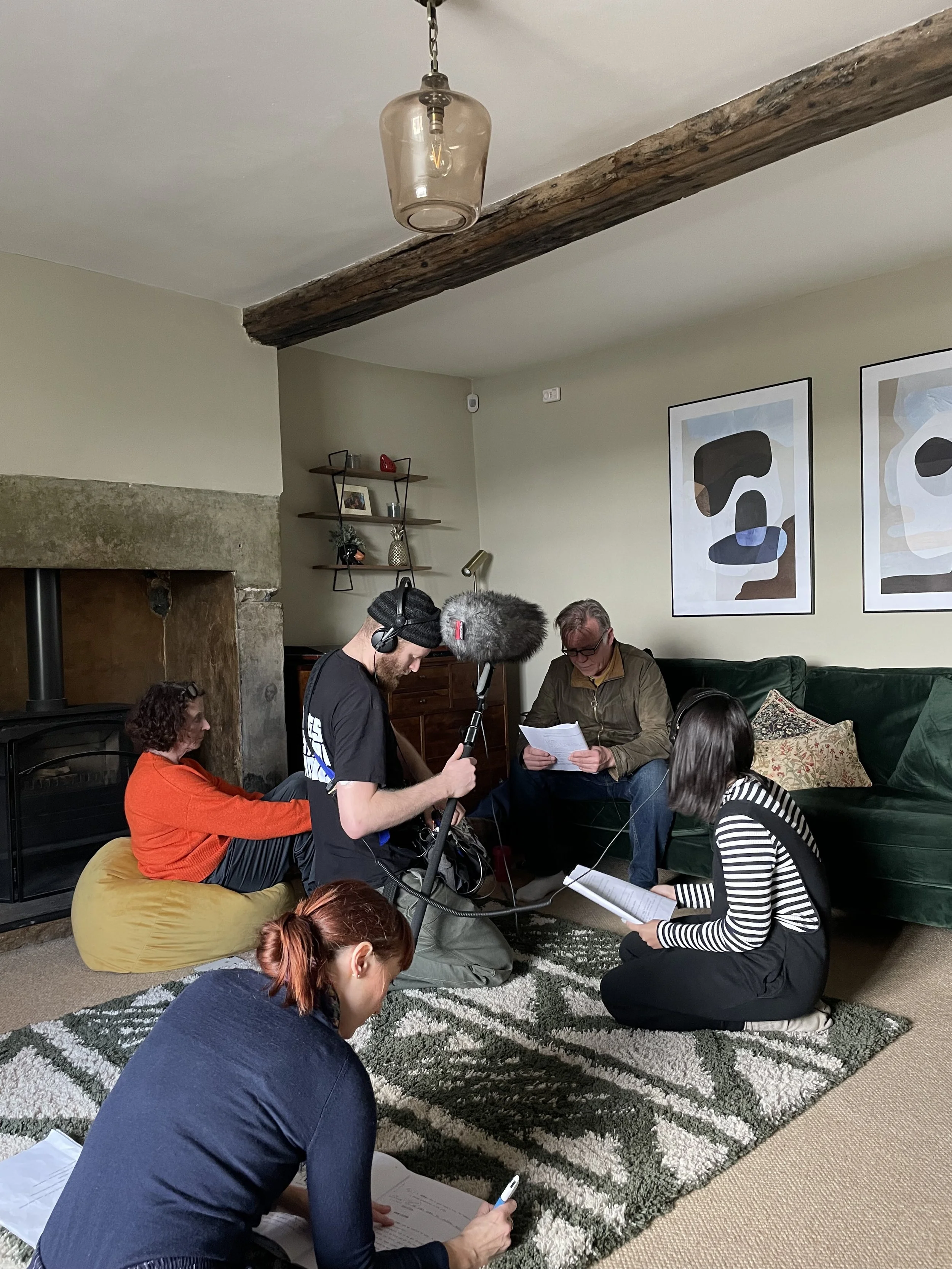 People in a living room participating in a film or audio recording session, some sitting on the floor, others standing and holding scripts and equipment.