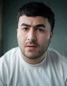 A man with dark hair and facial hair, wearing a white shirt, looking into the camera with a neutral expression.