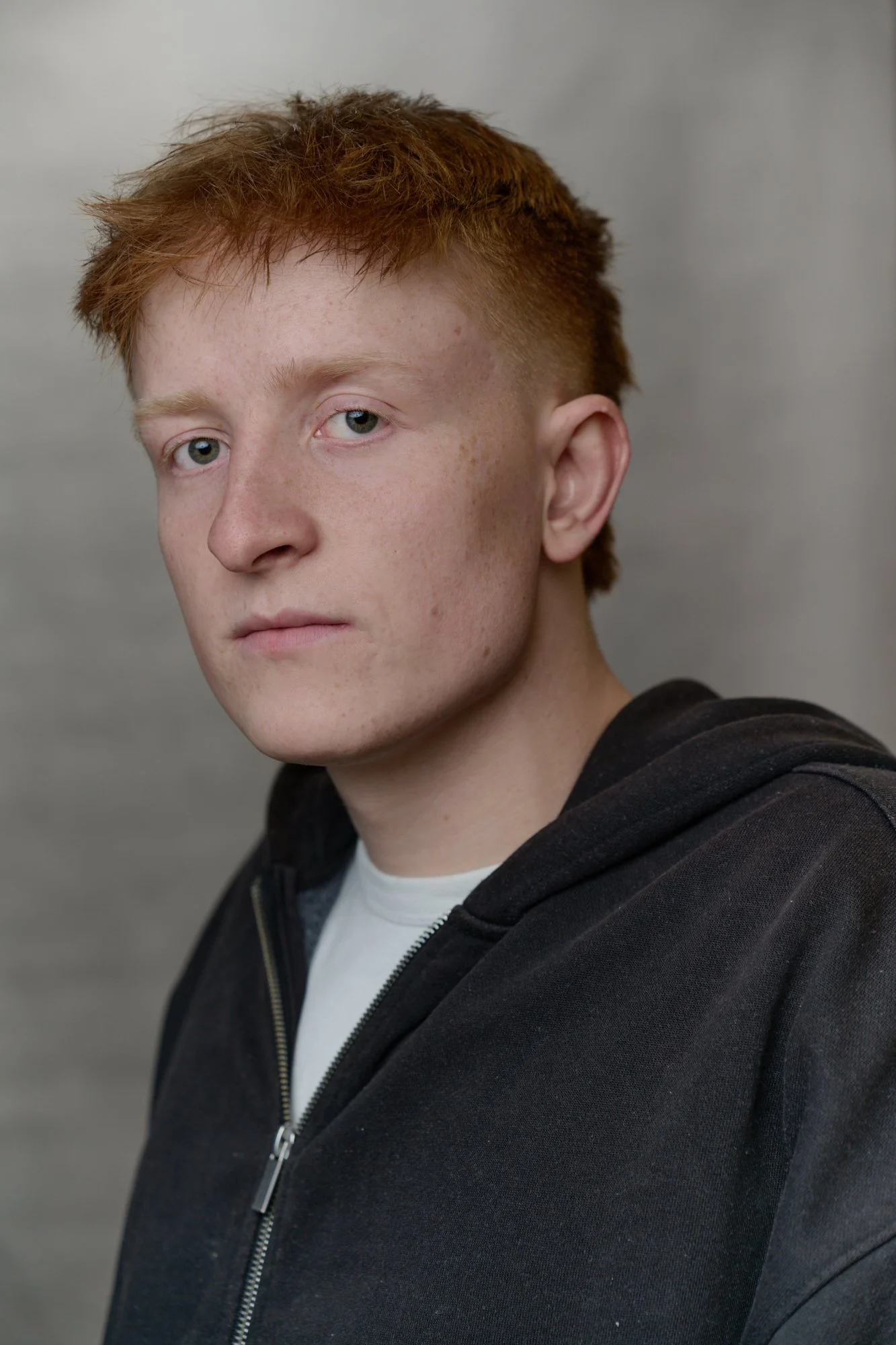 A young man with red hair and blue eyes wearing a white t-shirt and a black zip-up hoodie, looking at the camera against a plain gray background.