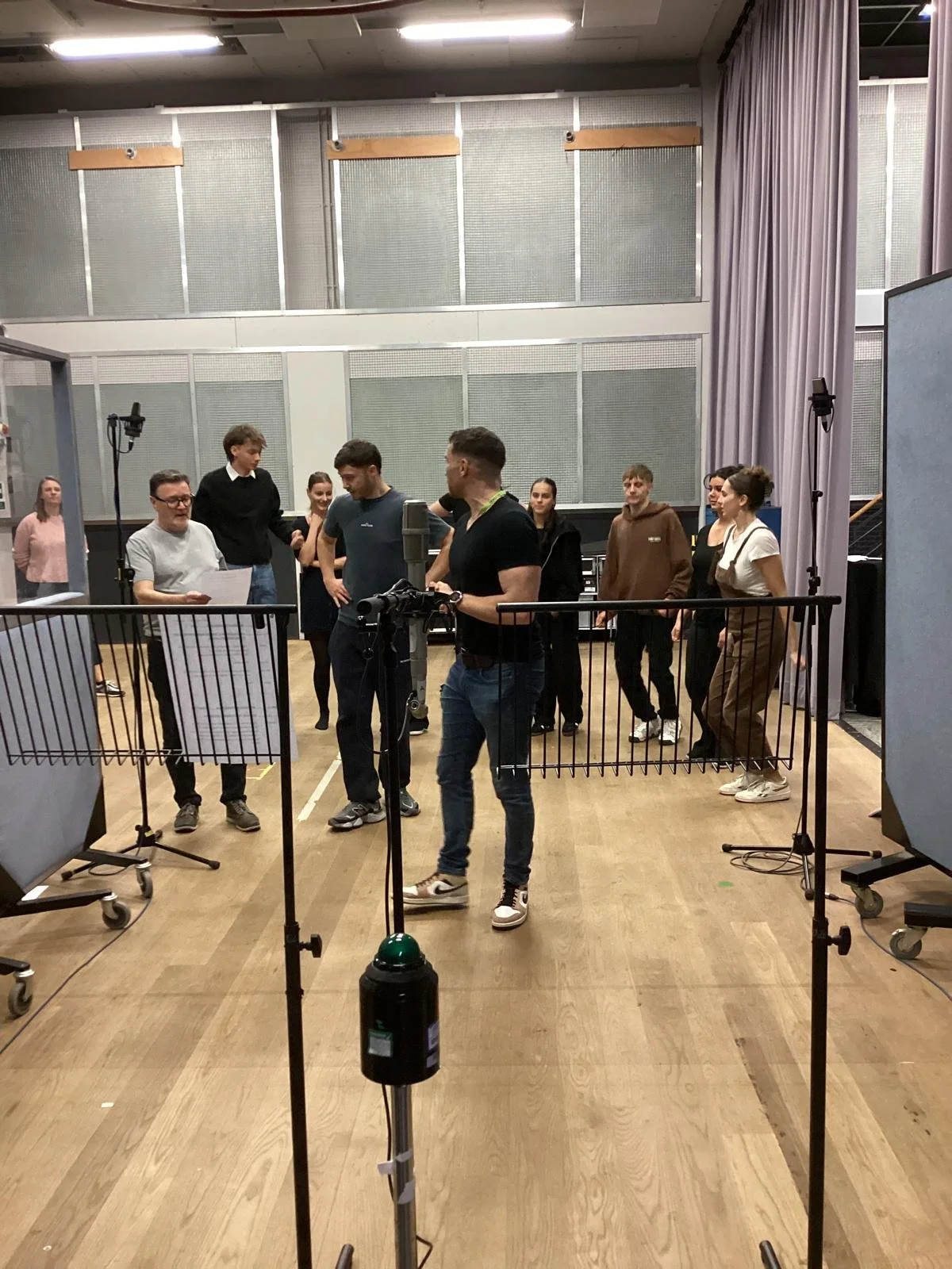 Group of people standing in a rehearsal space with microphones, audio equipment, and lighting, preparing for a recording or performance.