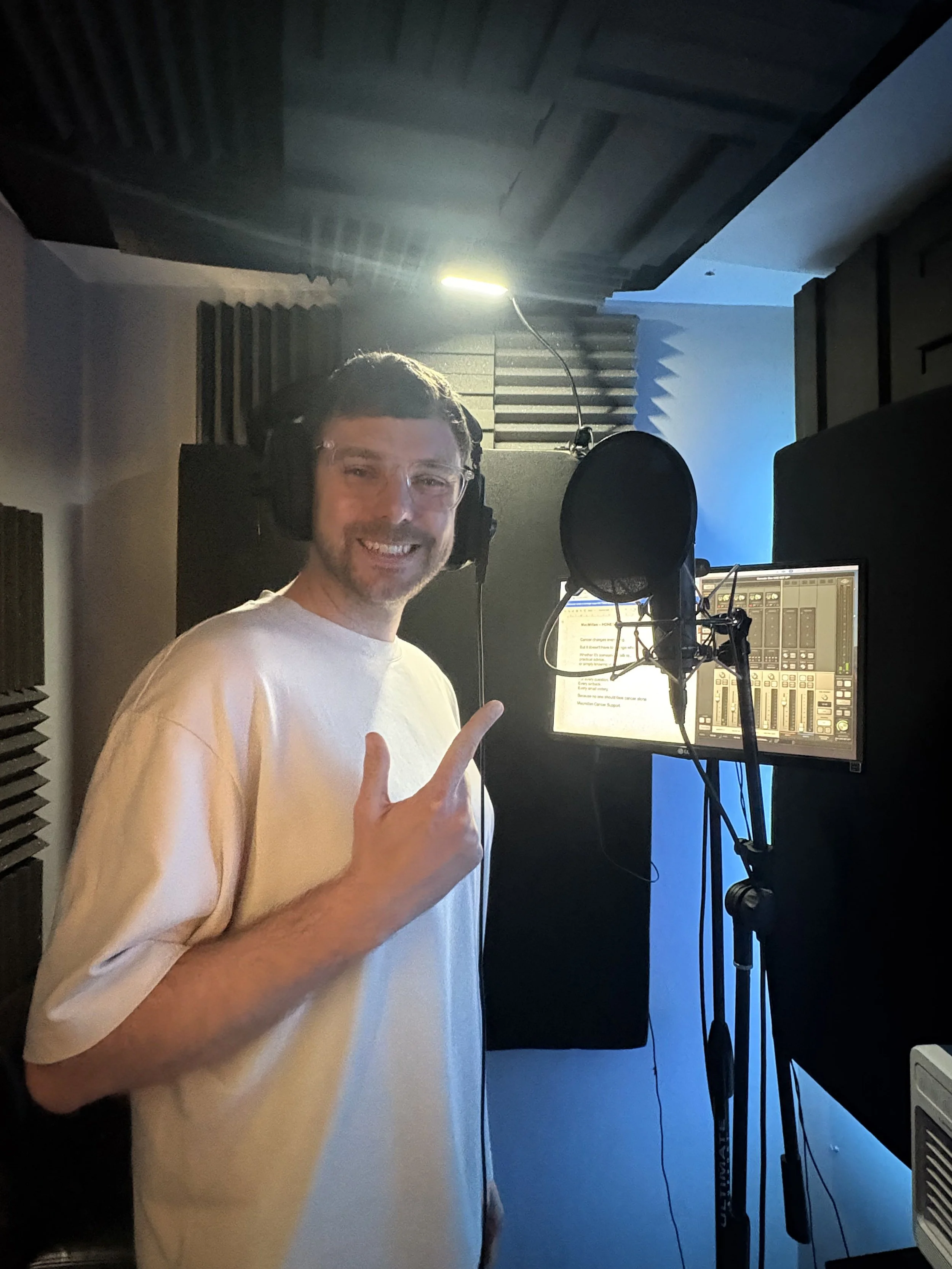 A man smiling in a recording studio, wearing headphones and clear glasses, standing in front of a microphone, with a computer and soundproofing foam in the background.