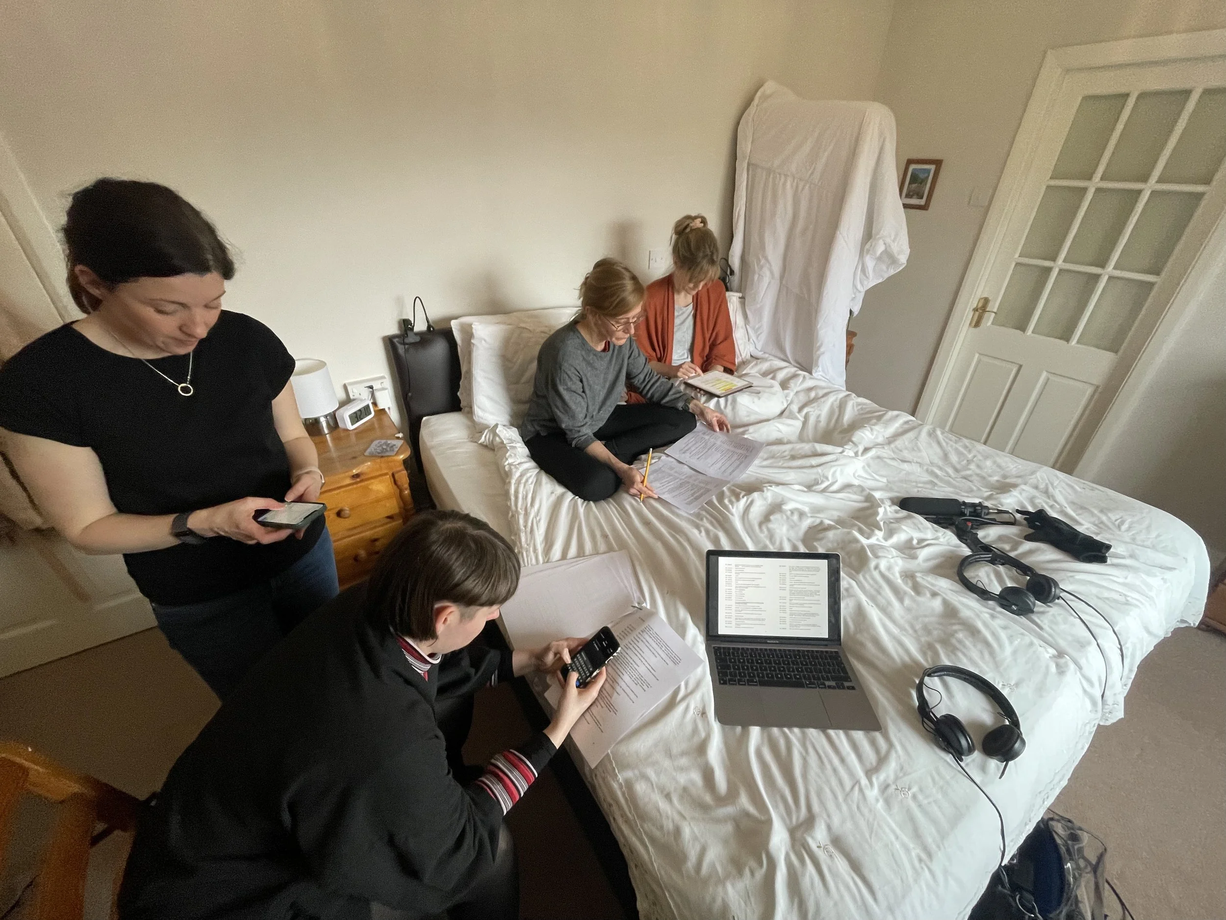 Five people in a bedroom with a bed, working with electronic devices, papers, and notebooks. One person is standing, looking at their phone, while others are seated or on the bed, engaging with papers and electronic gadgets.