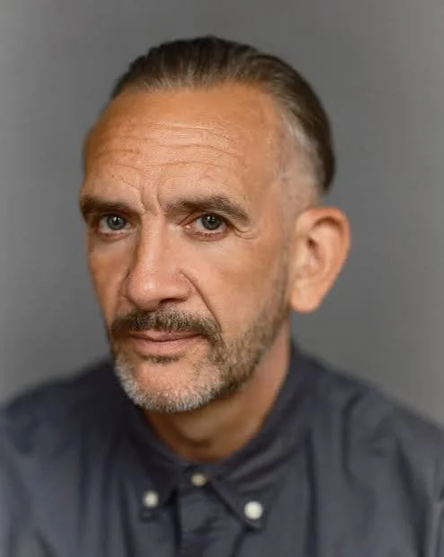 Close-up portrait of a middle-aged man with short, slicked-back gray hair, a beard, and wearing a dark collared shirt, against a plain gray background.