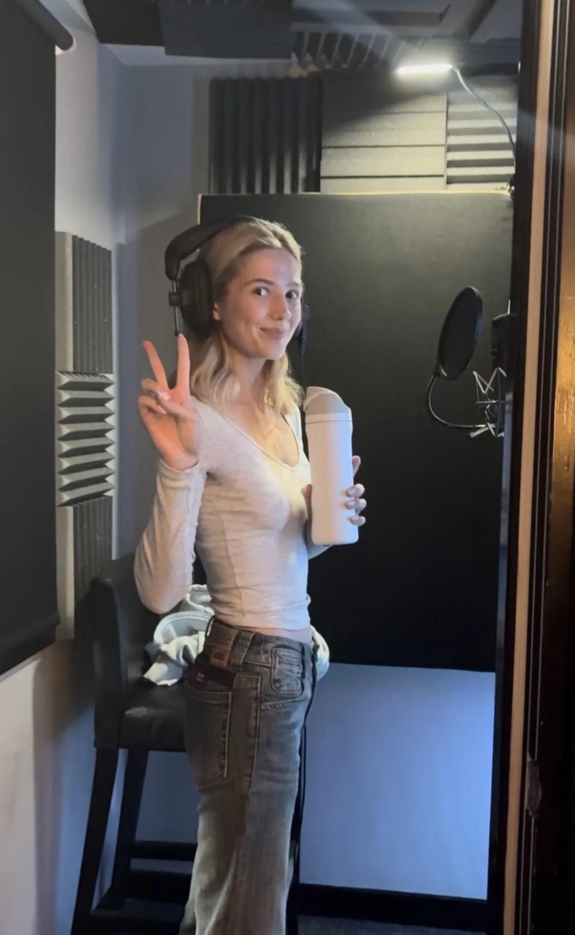 A young woman in a recording studio holding a water bottle and making a peace sign with her right hand.