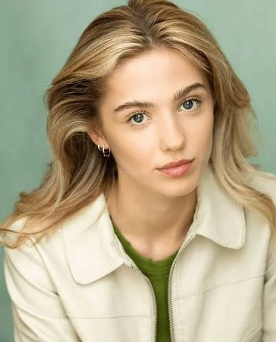 A young woman with blonde hair, blue eyes, and a neutral expression, wearing a beige jacket over a green top, against a light green background.