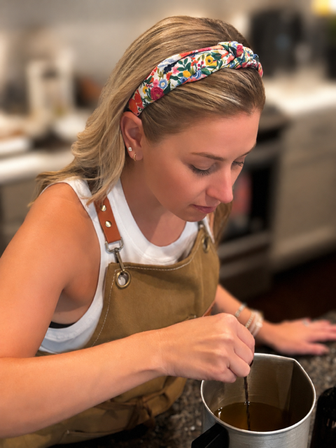 A young woman with blonde hair, wearing a colorful floral headband, a white sleeveless top, and a brown apron, is mixing a drink in a metal pitcher in a kitchen.