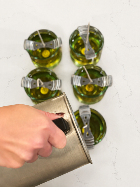 A hand pouring candle wax into clear glass candles with wicks from a metal melting pot.