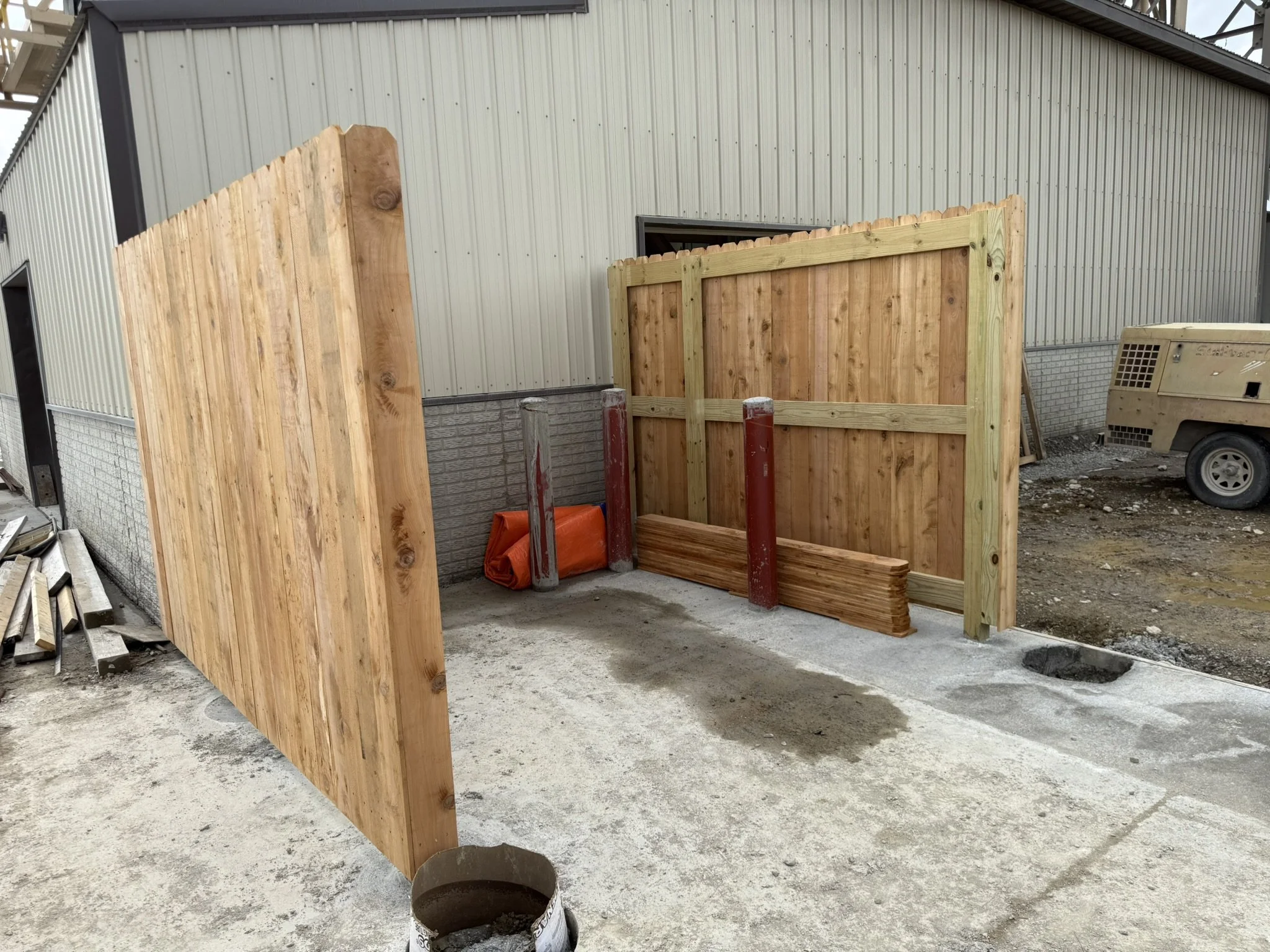 Construction site with wooden panels and a large metal building in the background. There are construction materials and a portable generator nearby.