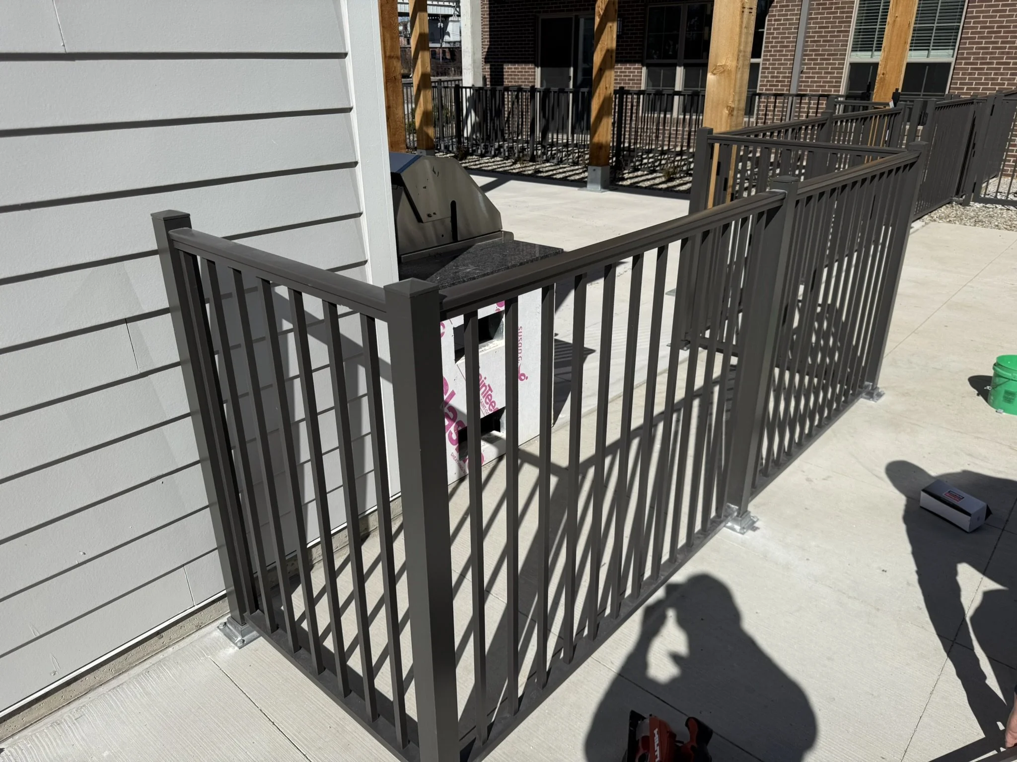 An outdoor deck area with a metal railing, a small grill, and part of a house with light-colored siding and brick walls in the background.