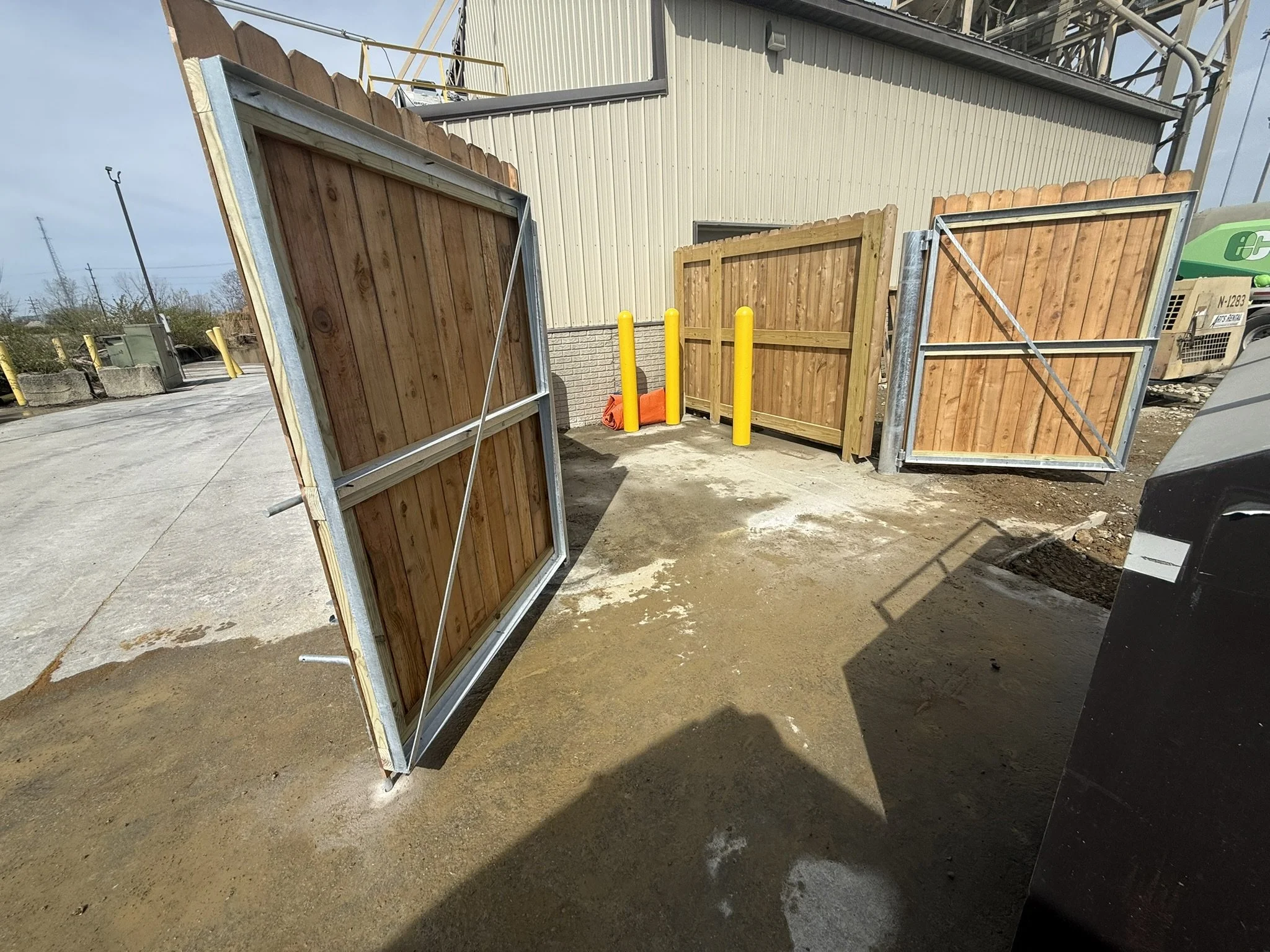 Open metal-framed gates with wooden panels at a commercial or industrial facility entrance. The gates are in the process of opening, revealing a building with metal siding and yellow safety bollards. There is a green utility vehicle nearby and power 