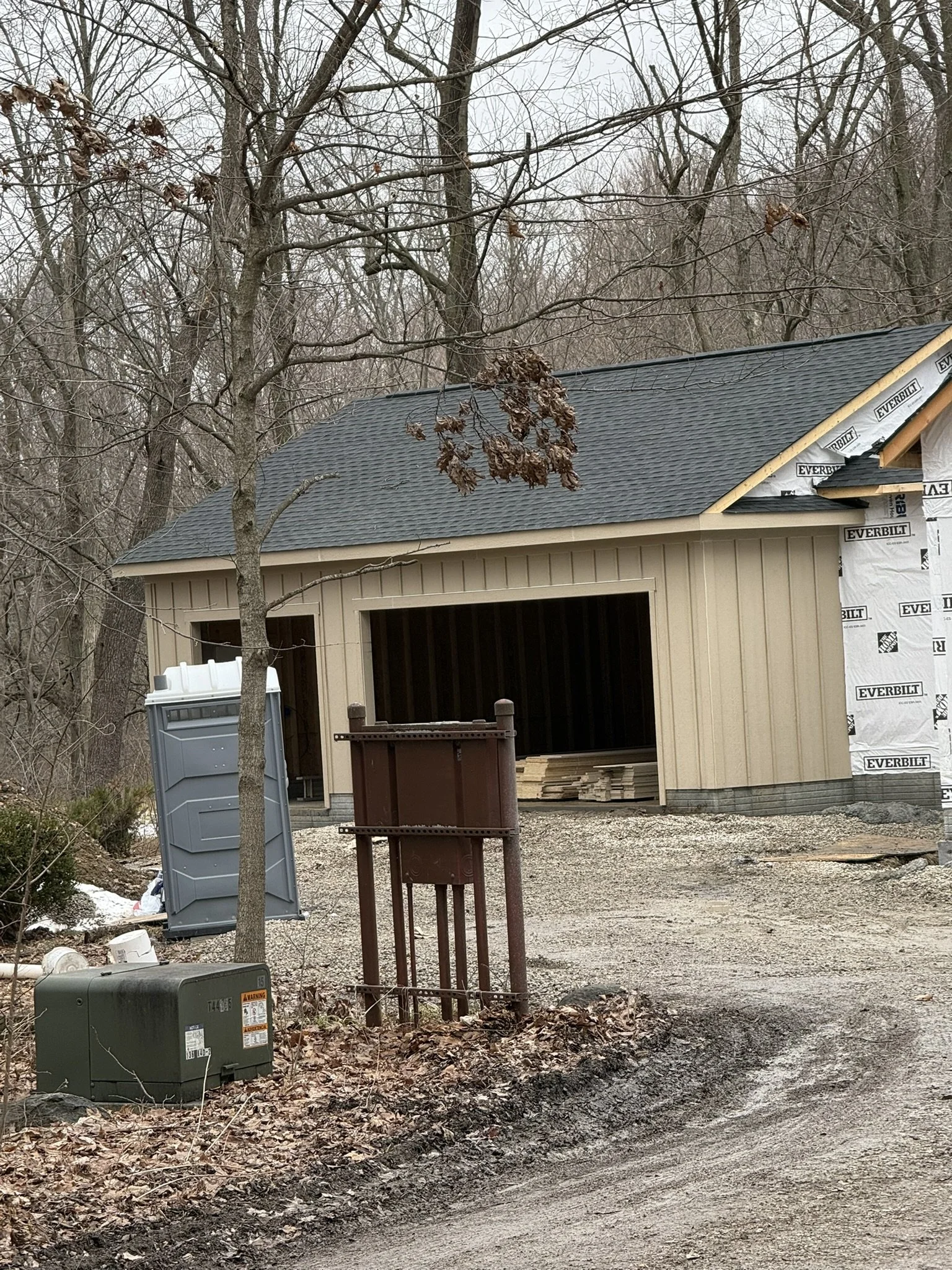 Construction site with a partially built garage, exposed framing, a black shingled roof, and construction materials around. A tree with brown leaves is in the foreground, and two trash bins are also visible.