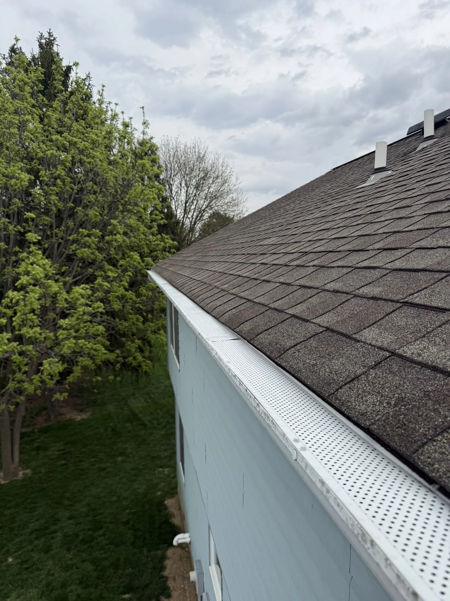 View of the side of a house with brown shingle roof and a blue wall, surrounded by green trees and grass, under a cloudy sky.