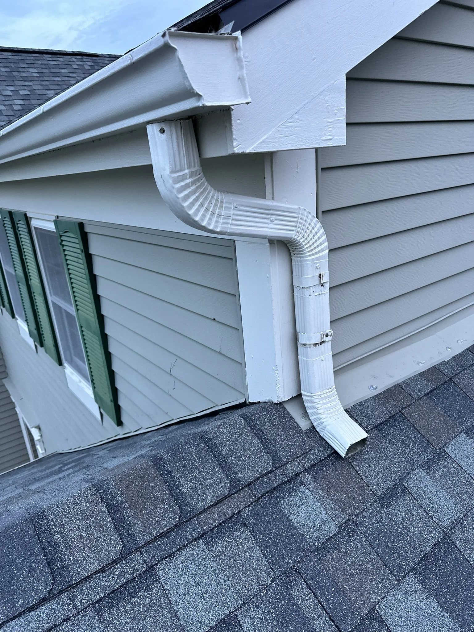 A close-up of a house's exterior corner, showing light grey vinyl siding, a white gutter system and downspout, and an asphalt shingle roof. The image is taken from an angle on the roof.