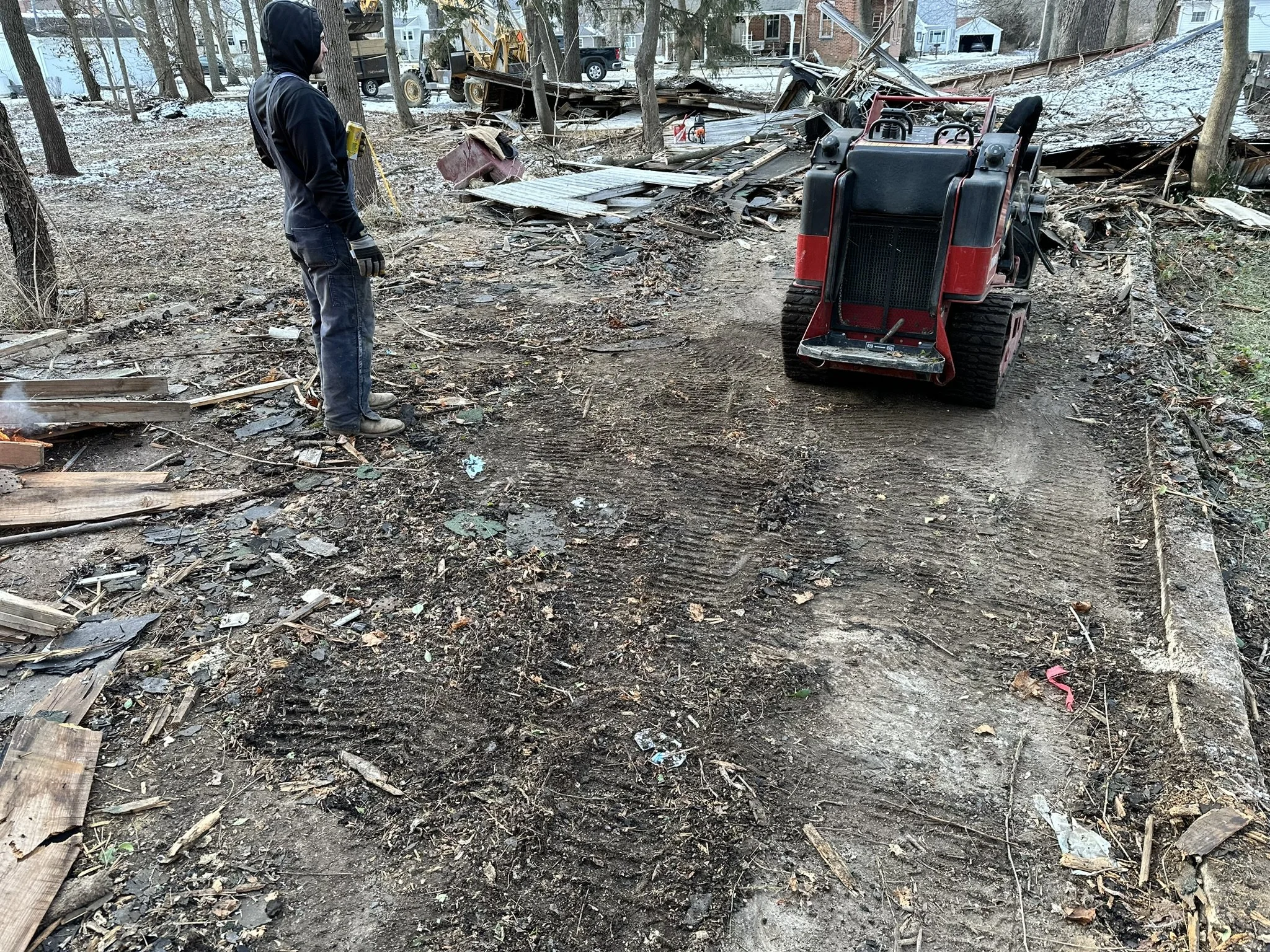 A person wearing a black hoodie and gloves looking at a damaged, destroyed area with debris, fallen trees, and a damaged vehicle in a wooded area after a storm or disaster.