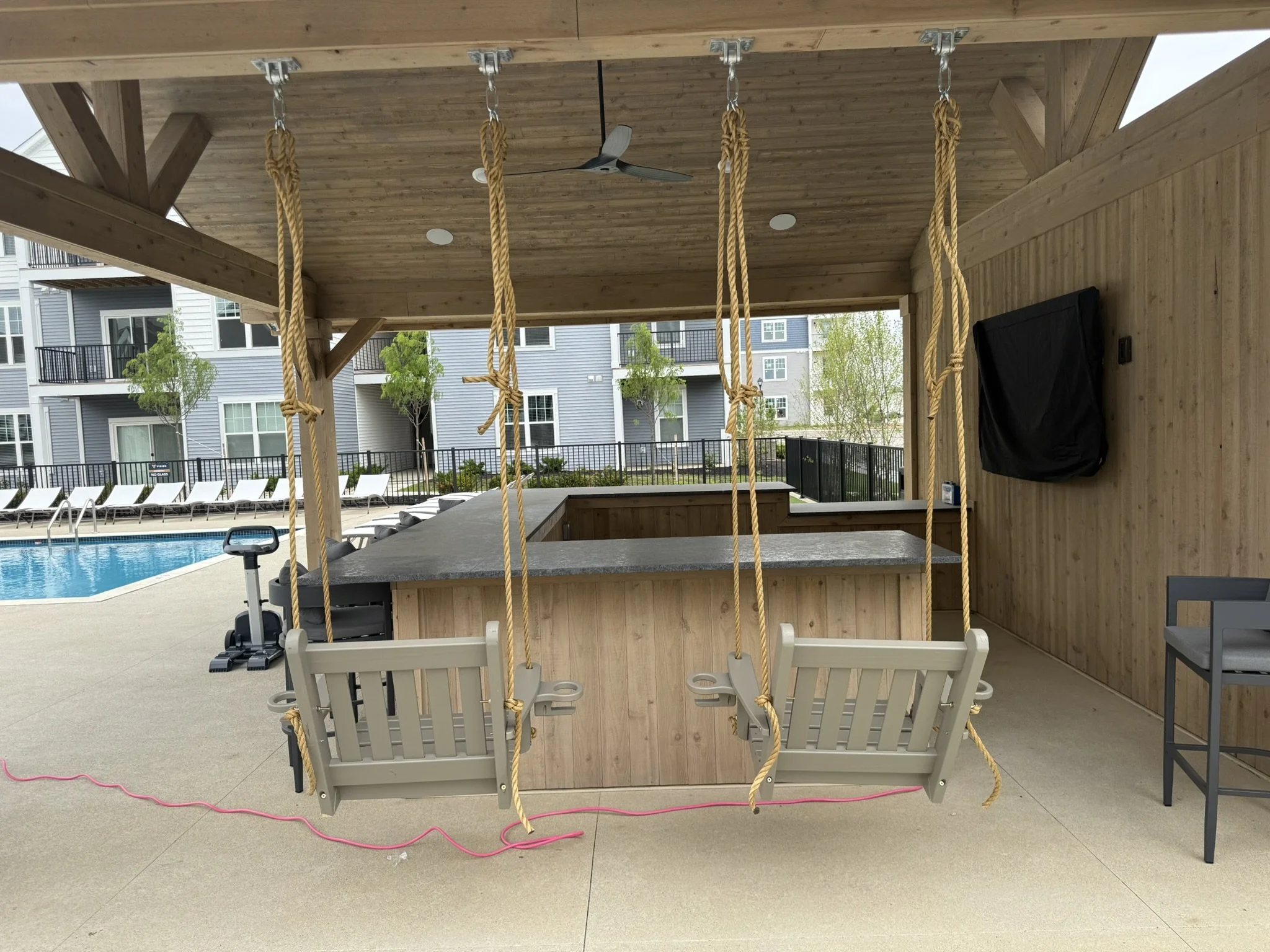 A backyard patio with a swimming pool, lounge chairs, and a wooden covered area. The covered area includes a ceiling fan, black flat-screen TV, and a wooden bar with rope swings hanging from the ceiling.