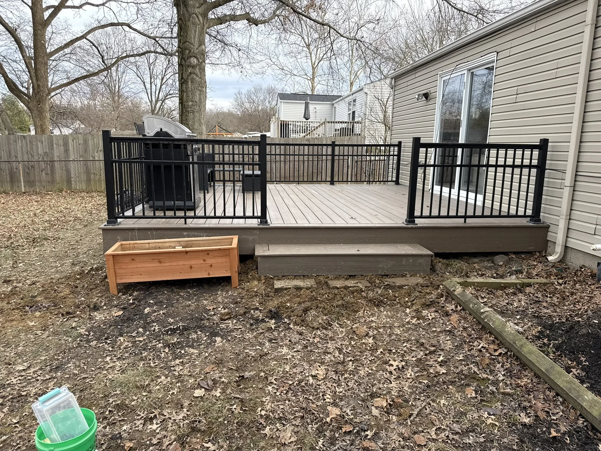 Backyard deck with a barbecue grill, outdoor seating, and a wooden planter box. Surrounding fence, trees, and outdoor furniture in the background.