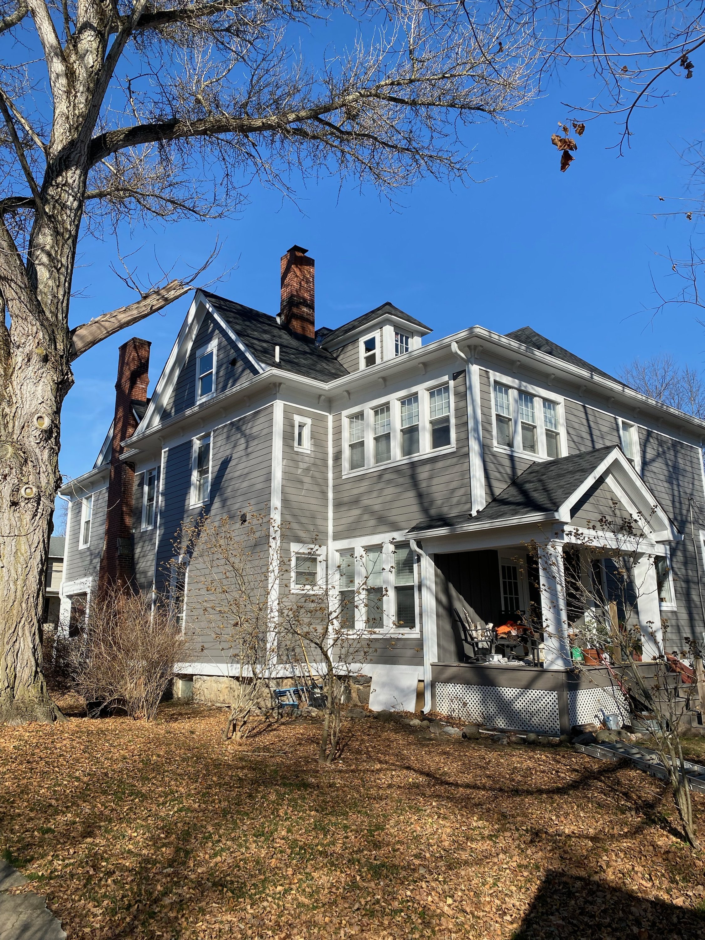 A large gray house with multiple windows, a porch with chairs, and a chimney, surrounded by leafless trees and a yard covered with fallen leaves, on a sunny day with a clear blue sky.
