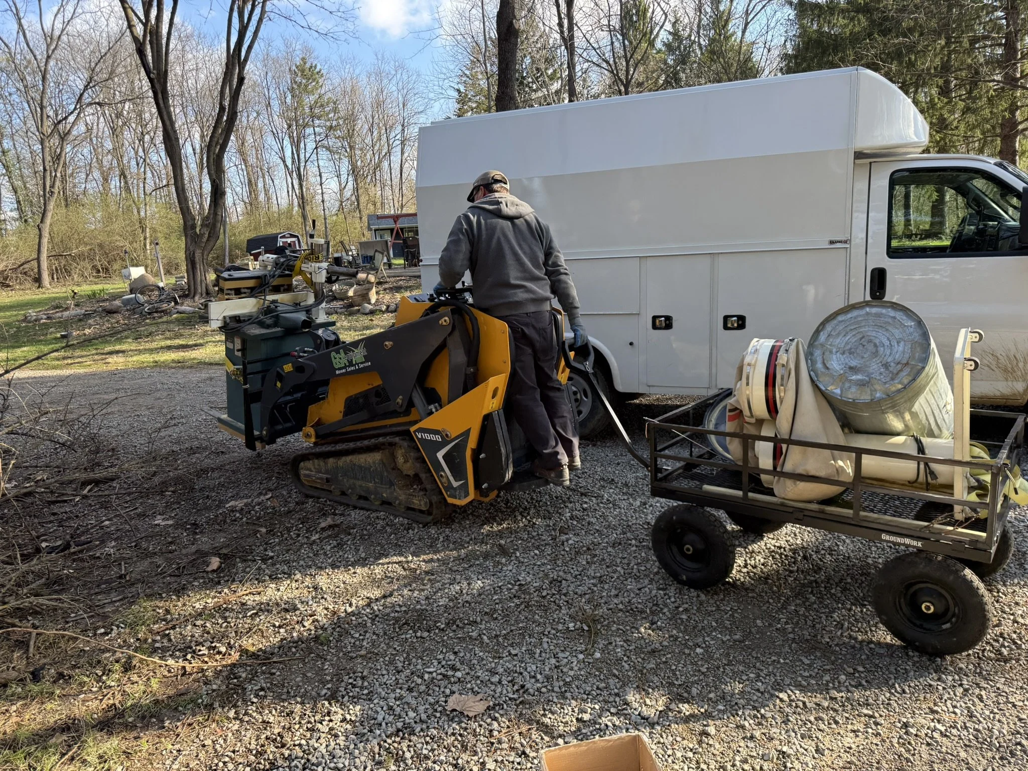 A person wearing a grey hoodie, black pants, overalls, and a cap working with a yellow and black mini excavator in an outdoor yard. There is a white enclosed truck, a trailer, and trees in the background.