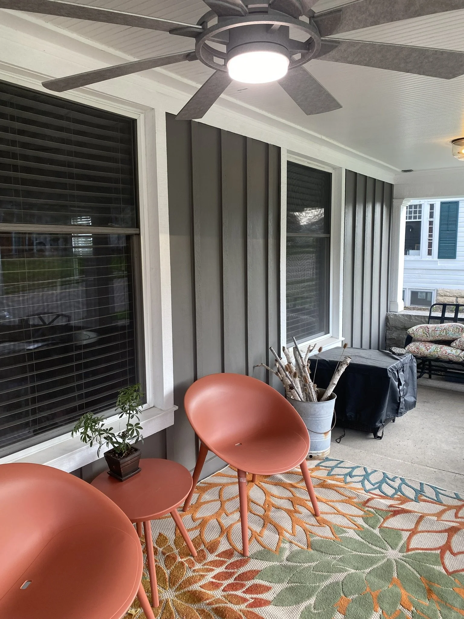 Covered outdoor patio with large windows, a ceiling fan, a small potted plant on an orange table, and patterned outdoor furniture. There is a colorful outdoor rug on the concrete floor.