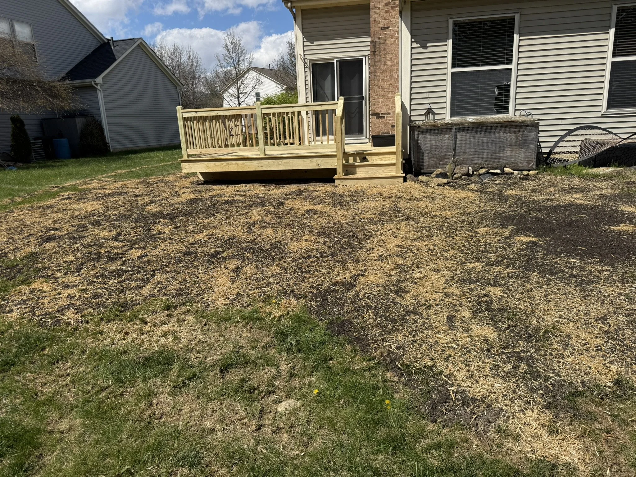 Backyard with new wooden deck attached to house, grass and black mulch area, neighboring houses in the background, partly cloudy sky.
