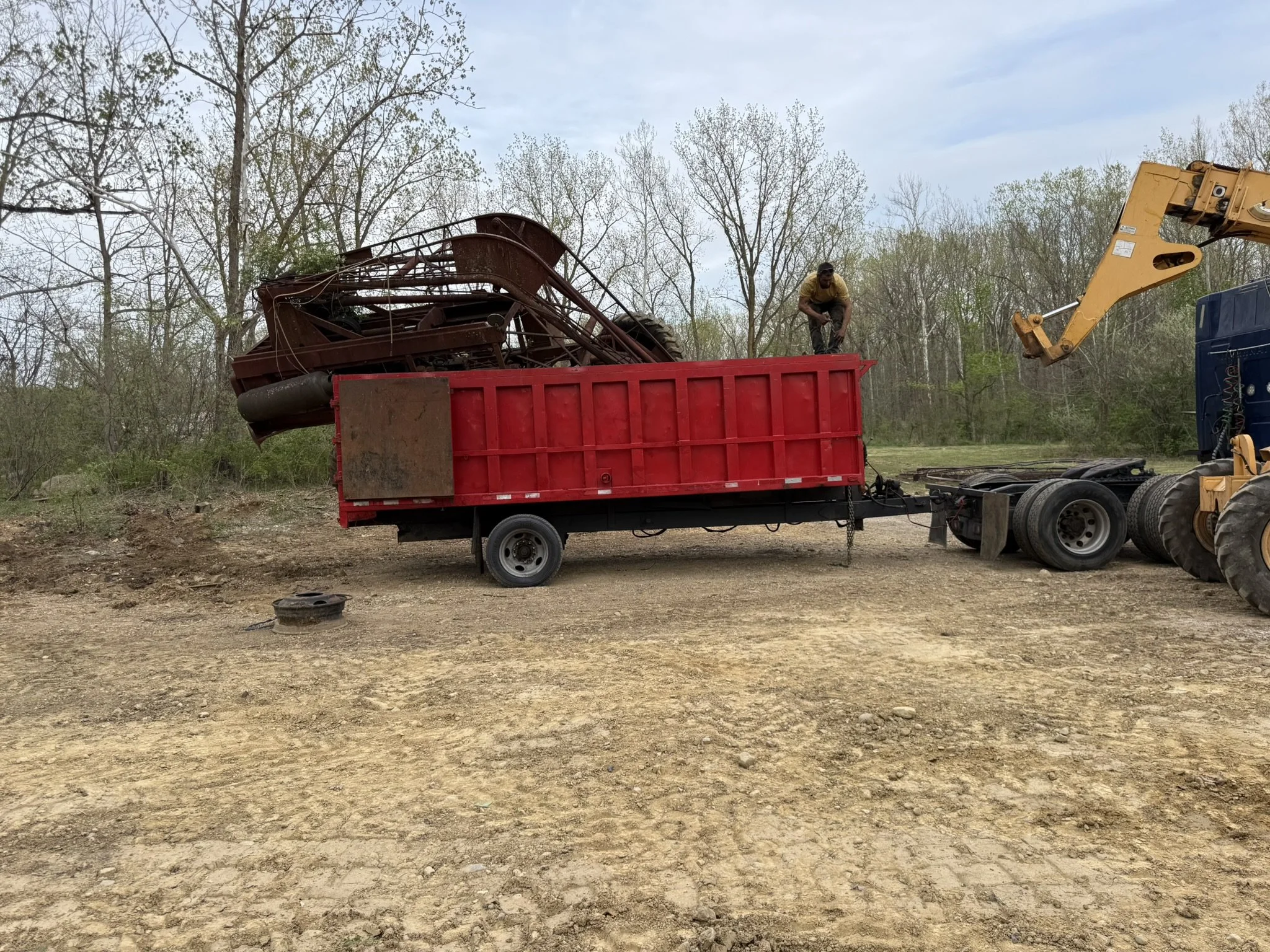 A person kneeling on top of a red truck bed, inspecting a large, rusted metal piece in a wooded area with a construction vehicle and trees in the background.