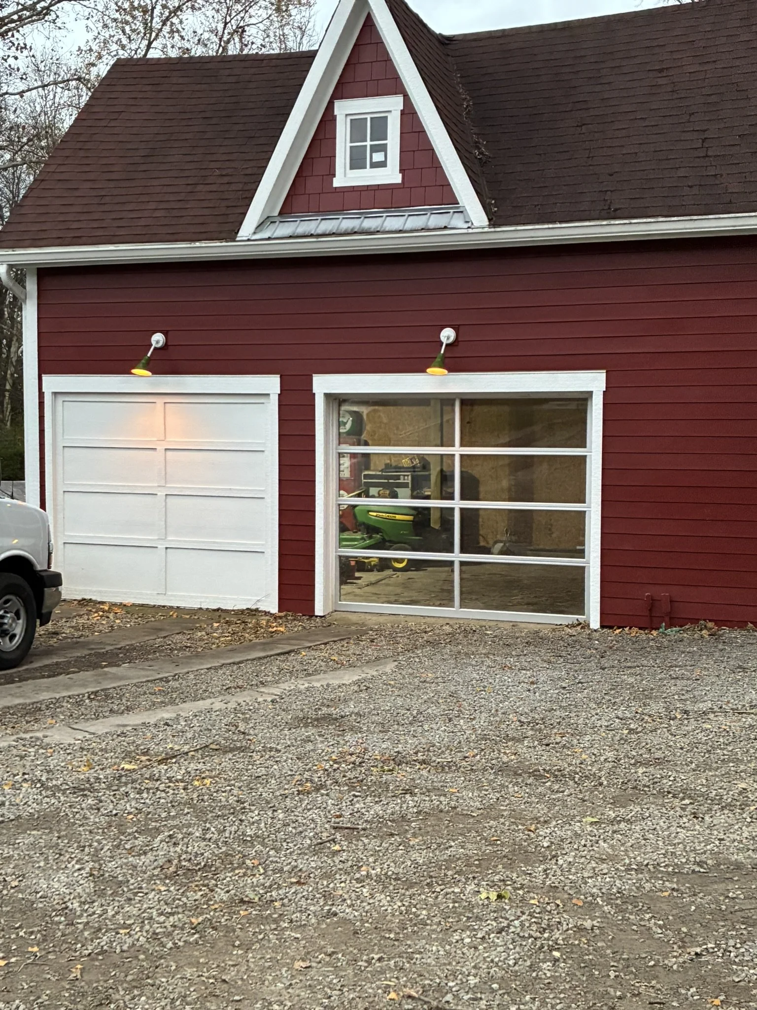 Red garage with a white door, a large window, and a small triangular window above, with construction or renovation in progress, tools and equipment visible inside through the window, and a gravel driveway in the front.