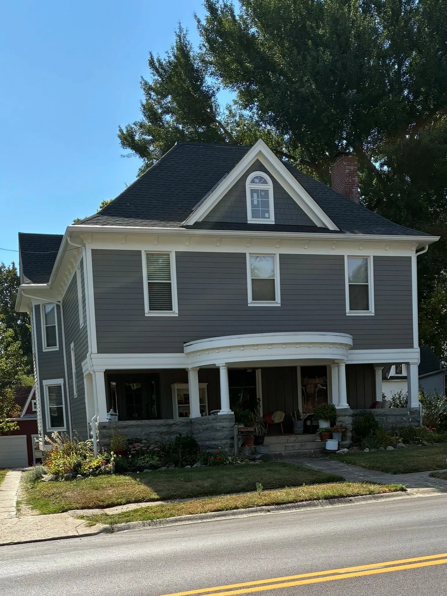 A large, two-story gray house with white trim, multiple windows, a covered porch, and a steep-pitched roof with an attic window. The house is surrounded by a garden with potted plants and flowers, and there is a sidewalk and street in the foreground. Tall trees are behind the house, and a blue sky is visible above.