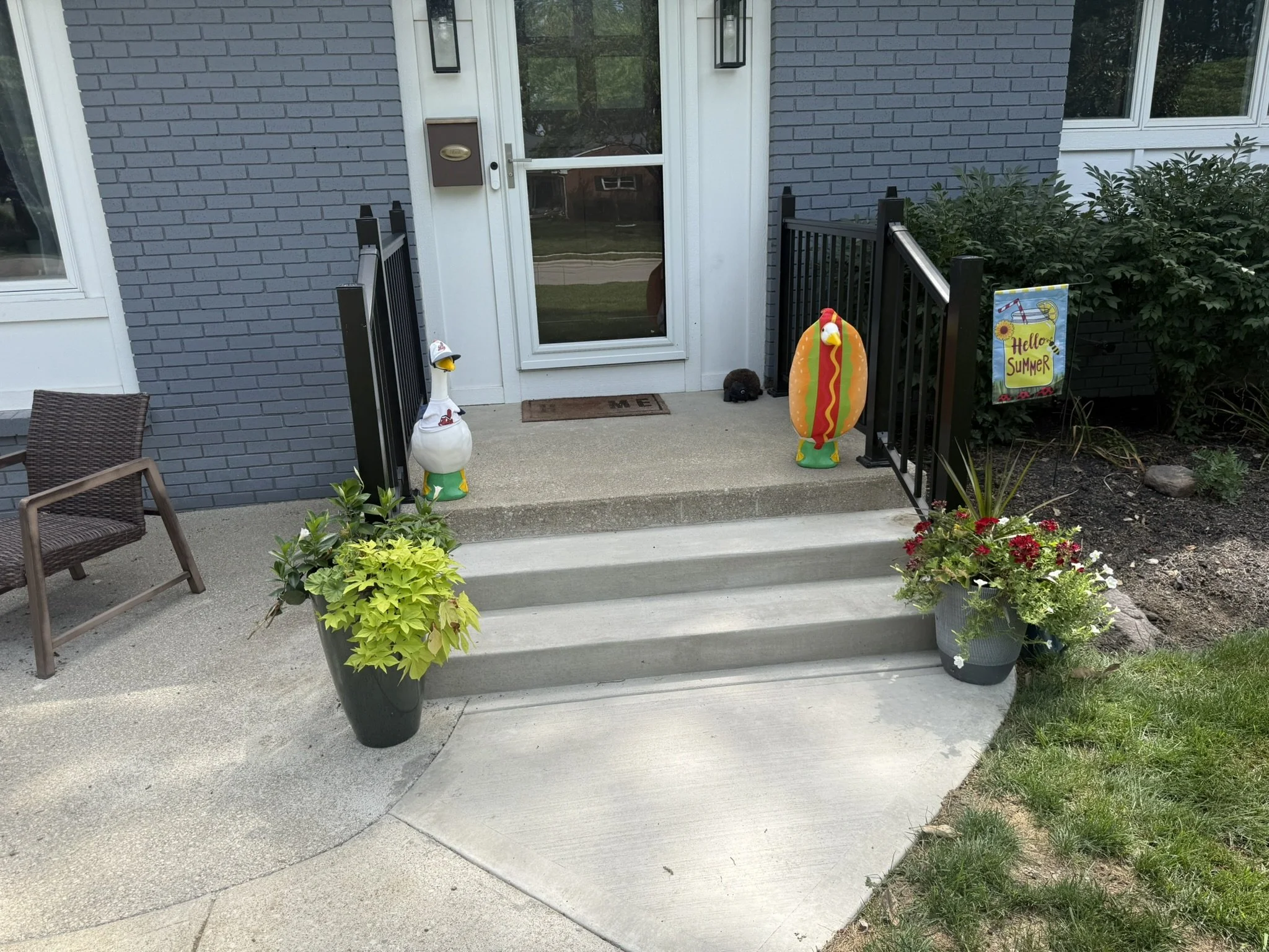 View of a house front porch with a gray brick wall, white door with a glass window, black railing, potted plants, a decorative flamingo, a hot dog inflatable, and a 'Hello Summer' sign, with plants and grass nearby.