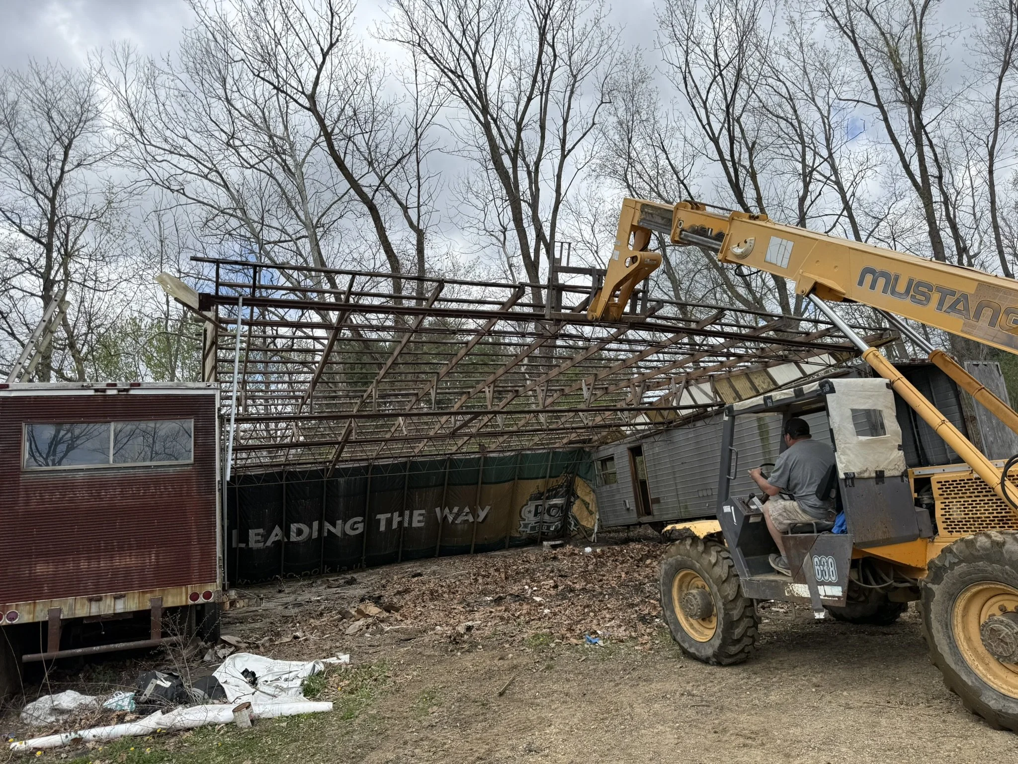 Construction worker using a yellow telescopic crane to lift a metal framework under leafless trees amidst construction debris and equipment.