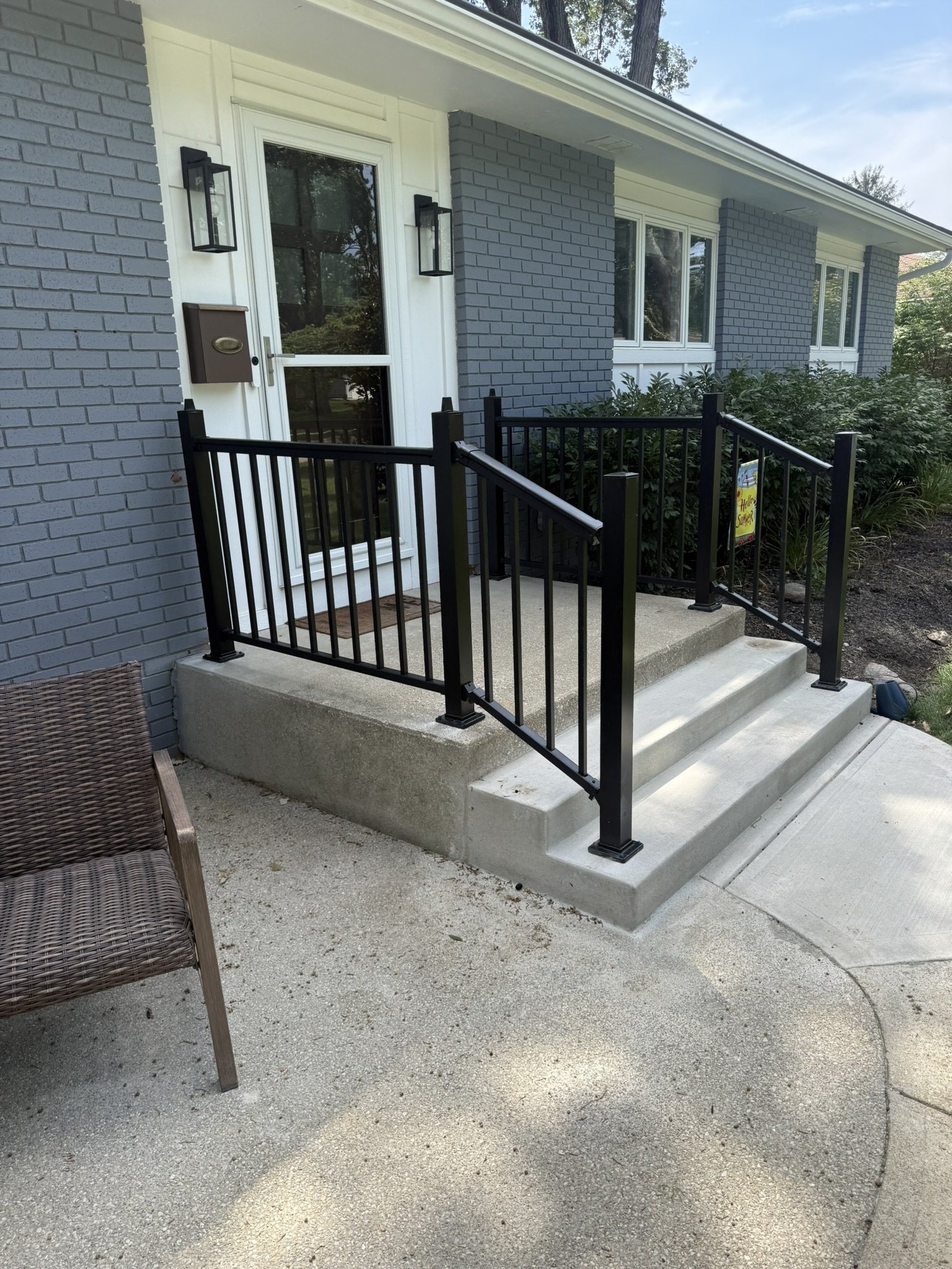 A brick house entrance with a glass door, surrounded by bushes, with a black metal railing on concrete steps leading up to the door. There are two wall-mounted lanterns beside the door, a mailbox, and a Welcome Summer sign near the bushes.