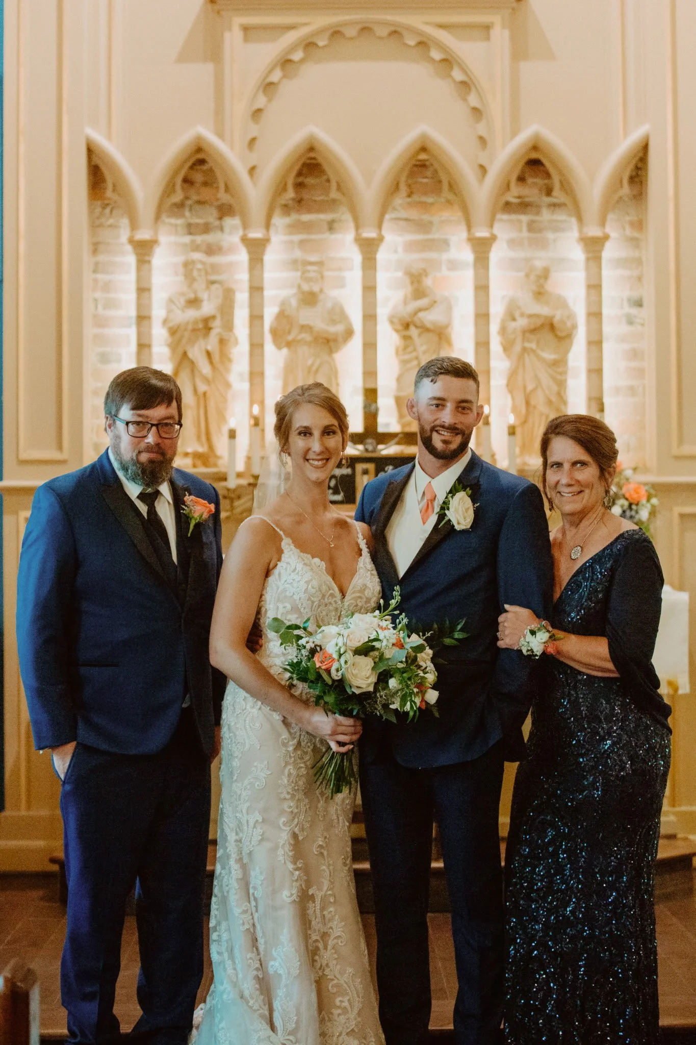 A wedding reception with a bride and groom in the center, flanked by two women and a man, standing inside a church with religious statues in the background.