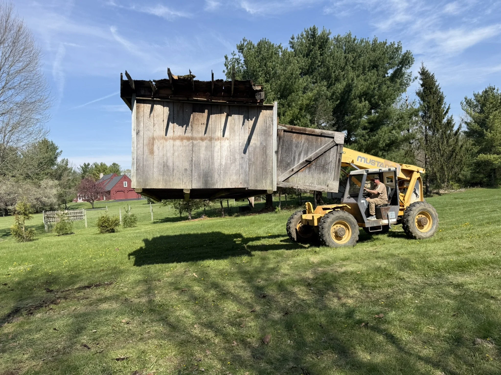A person operating a yellow John Deere backhoe loader on a grassy lawn, lifting a weathered wooden shed with a deteriorated roof, illuminated by clear blue skies.