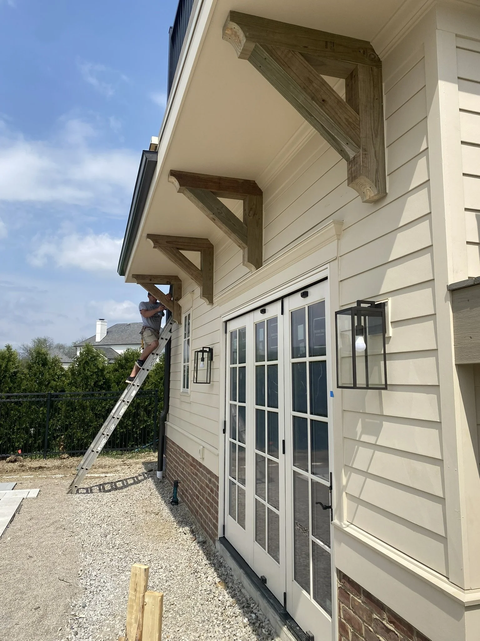 Workers installing wooden brackets under the roof overhang of a beige house exterior, with French doors and decorative black lantern-style lights.