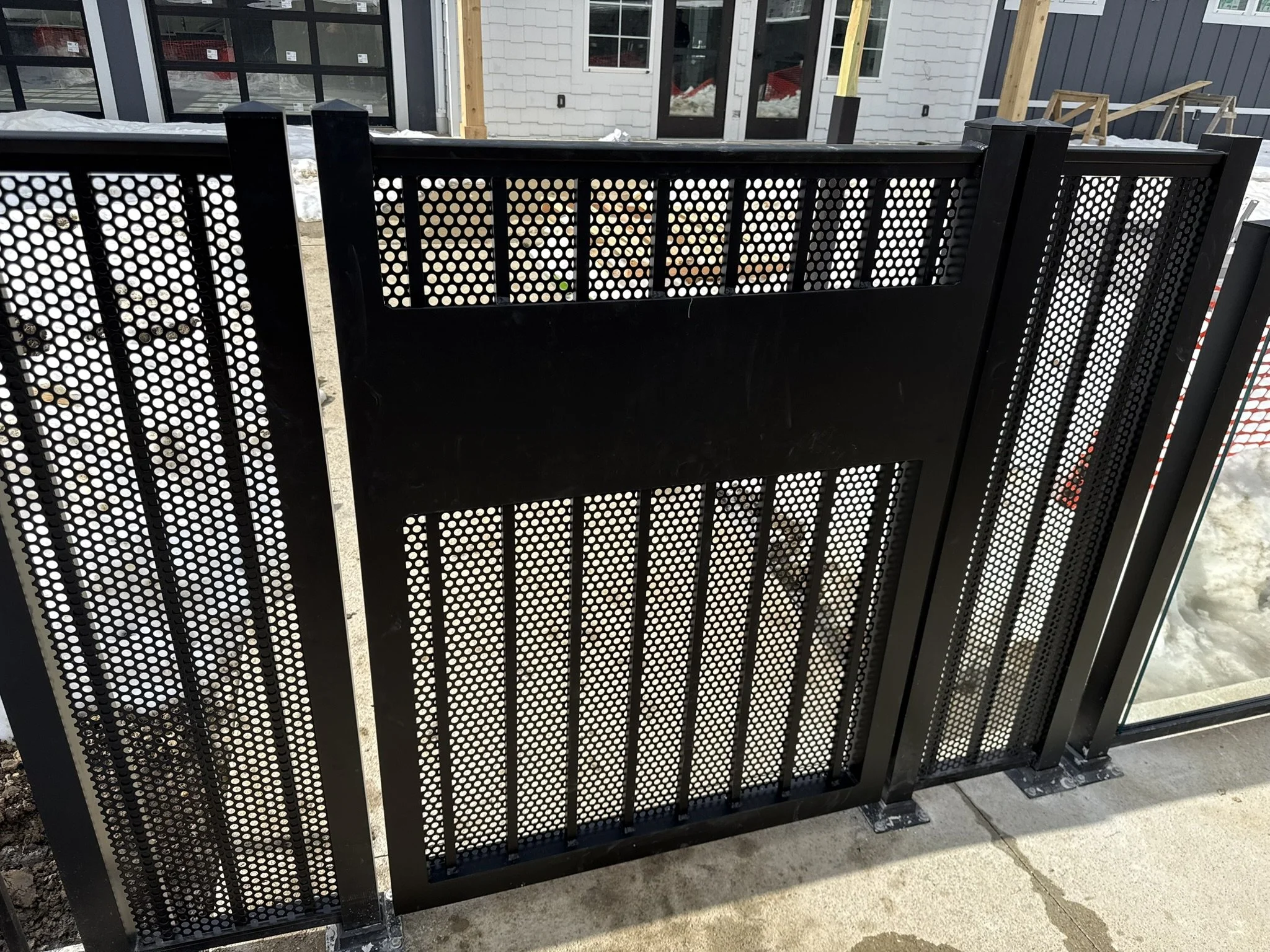 Black metal patio fence with circular perforations, installed on a concrete surface in front of a house under construction. The house has a gray siding exterior, a door with glass panes, and a partially completed porch with wooden framing.