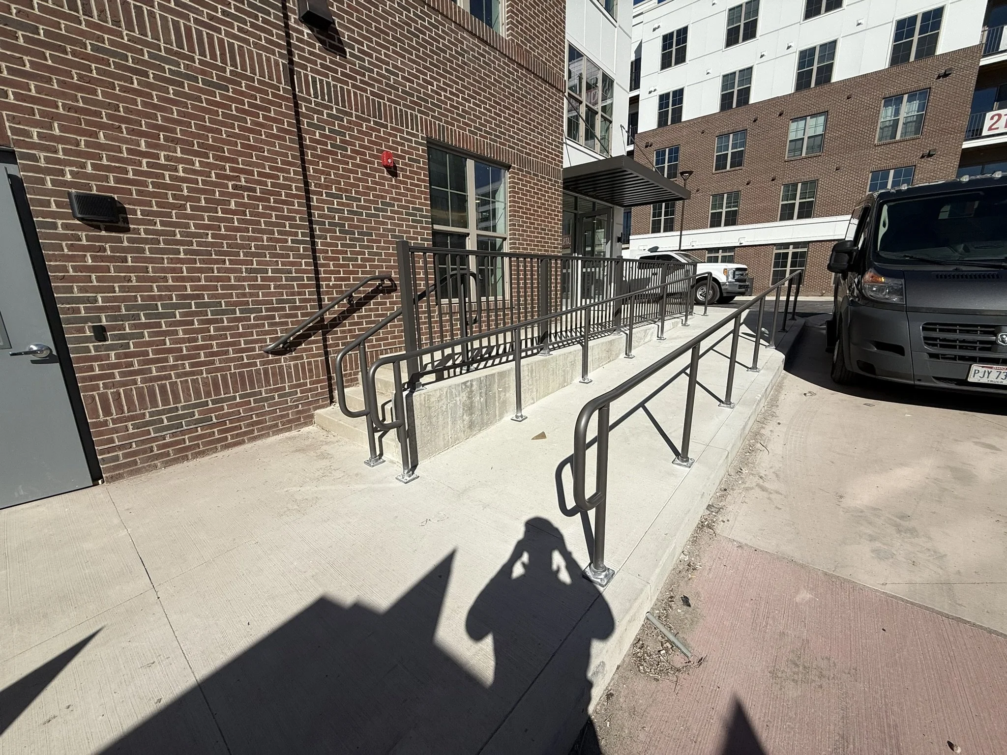View of the exterior of a brick apartment building with large windows, a glass door entrance, a metal awning, and a wheelchair accessible ramp. There are parked vehicles, including a gray van and a white pickup truck, on the sidewalk.