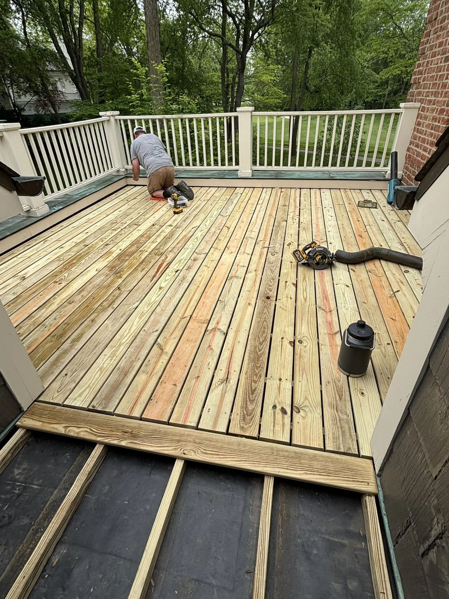 A person installing a wooden deck, with tools and partially completed floorboards, on a backyard patio surrounded by trees and a brick wall.