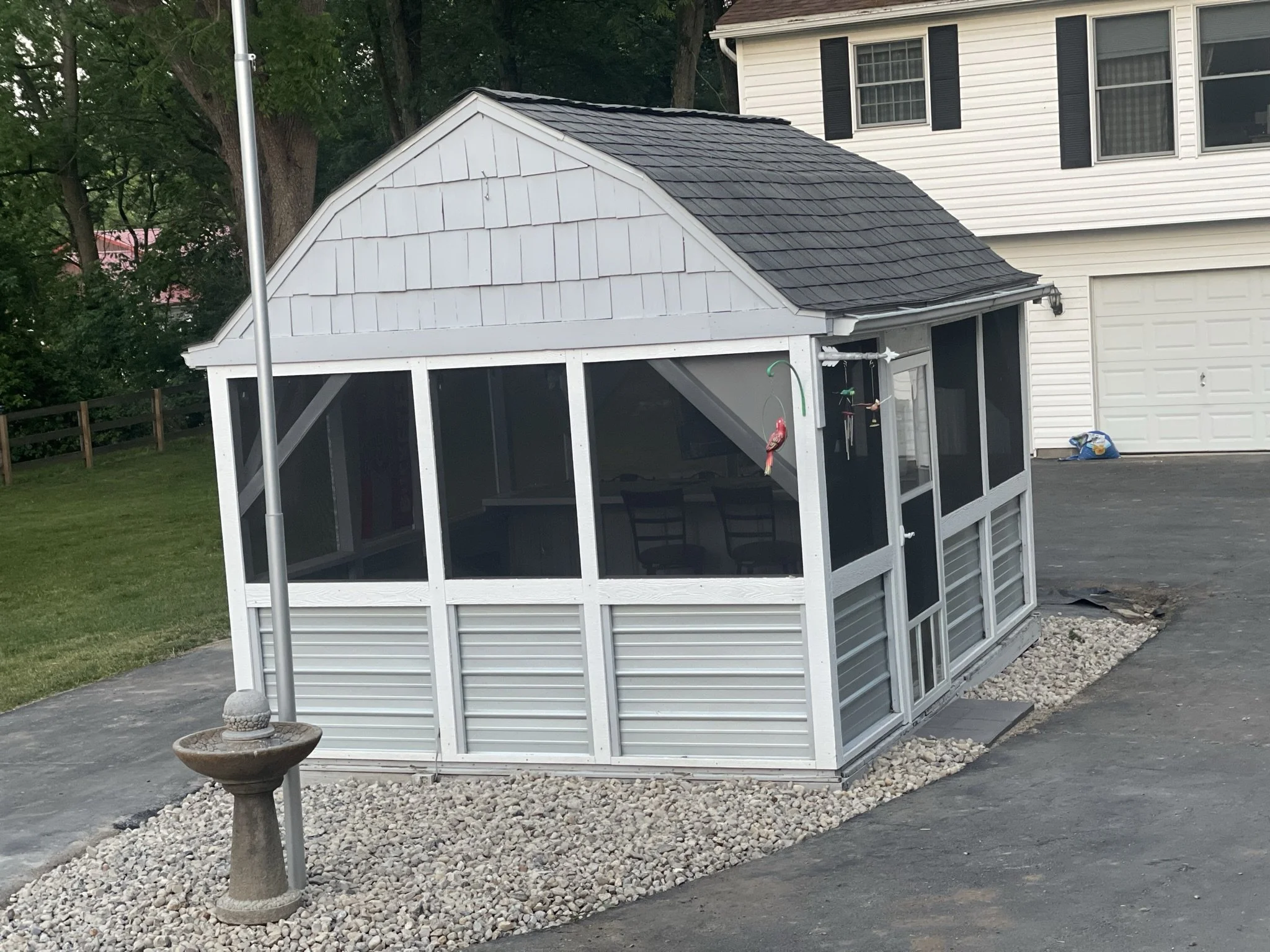 A backyard scene featuring a white screened gazebo with a dark shingled roof, a small fountain with a bird decorative element, and a house with white siding, black shutters, and a garage door. There are trees and a grassy area in the background.