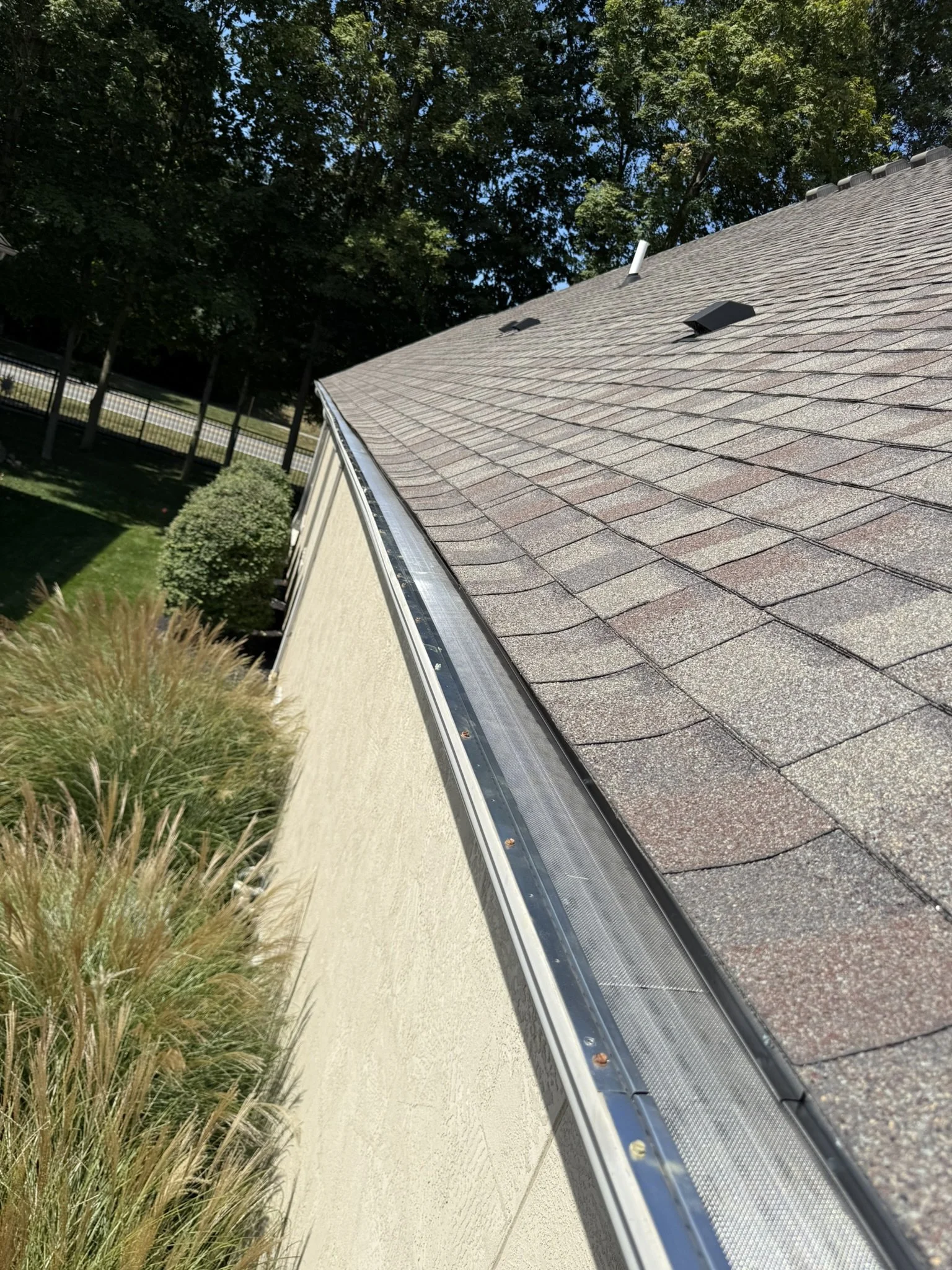 Residential house roof with asphalt shingles, plumbing vent, vents, and solar panel, surrounded by trees and a fence.